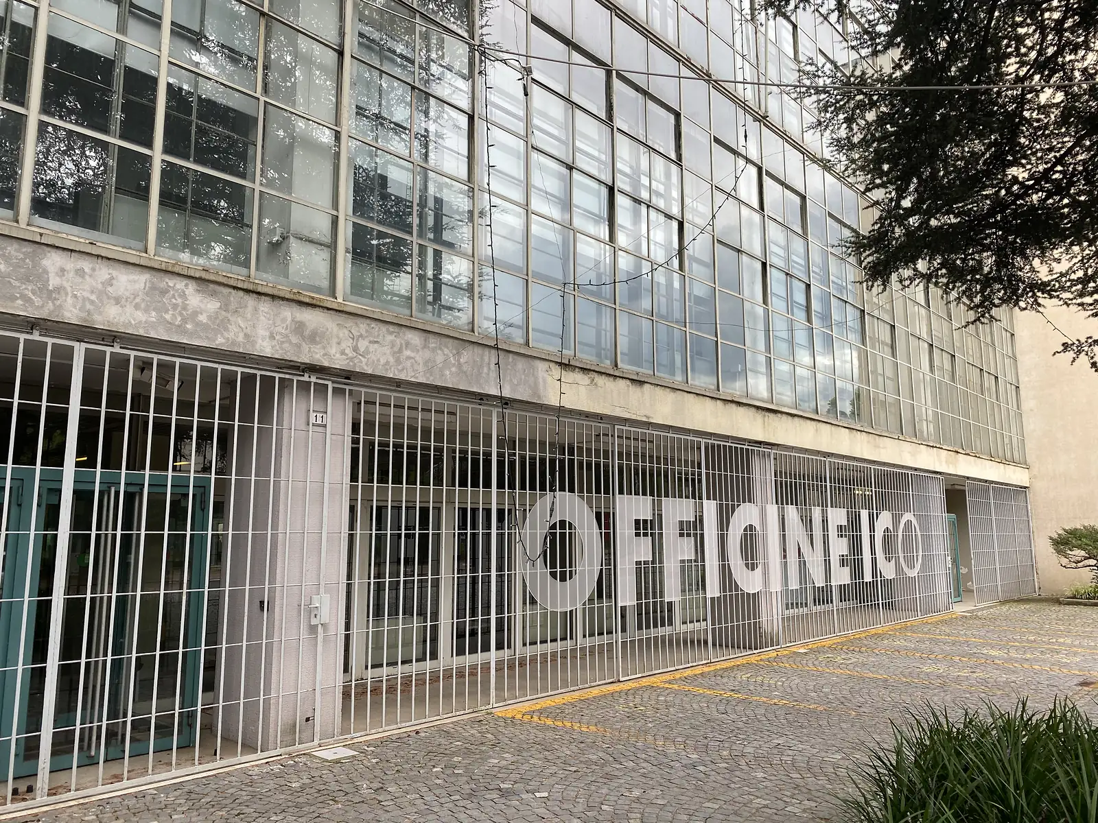A barred glass shopfront reading "Officine ICO" beneath the tiered glass curtain wall of a former factory building, a tree overhanging from the right.