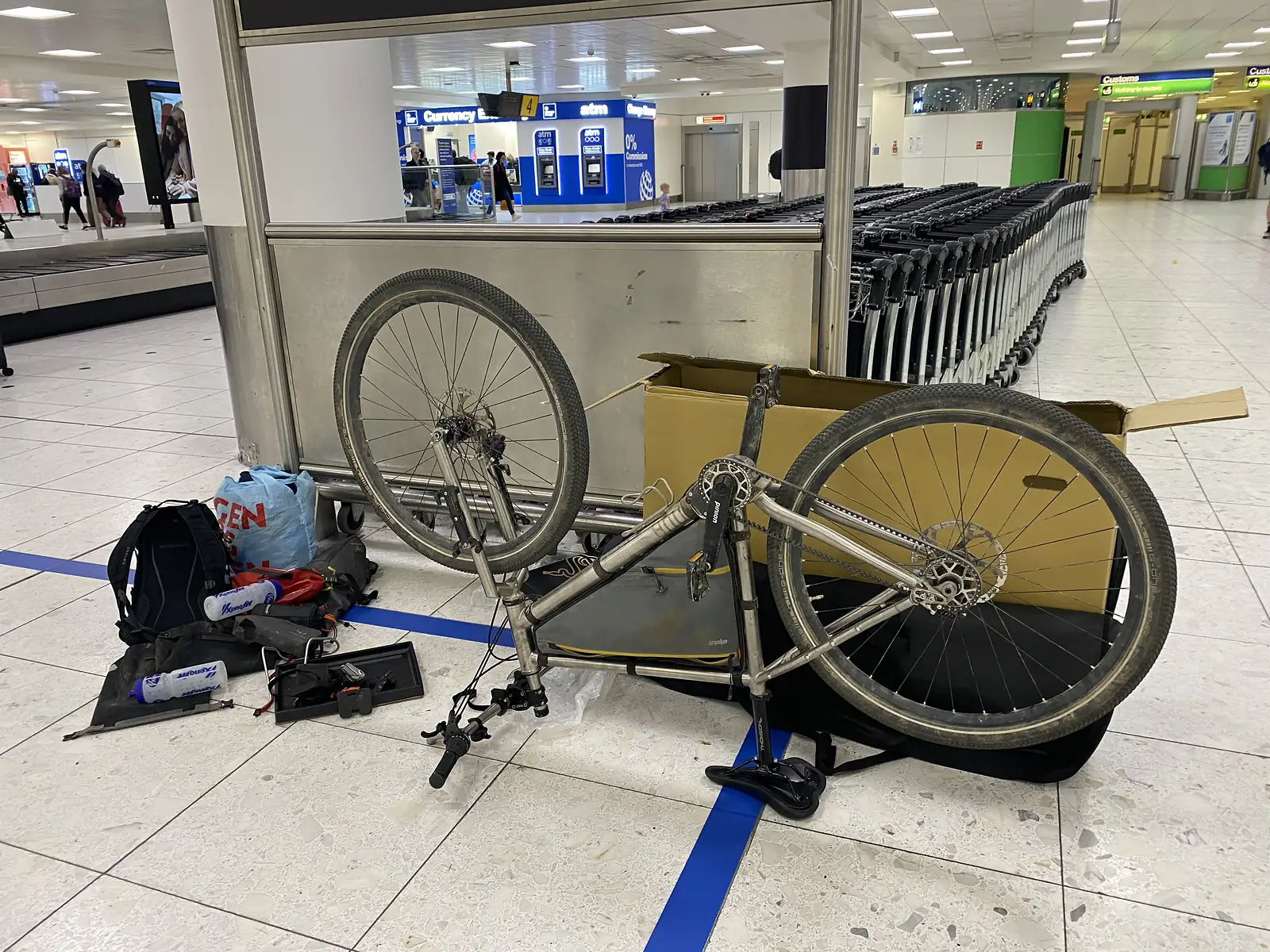 A bike frame, wheels and tools laid out on an airport arrivals floor beside luggage trolleys, with currency exchange desks behind.