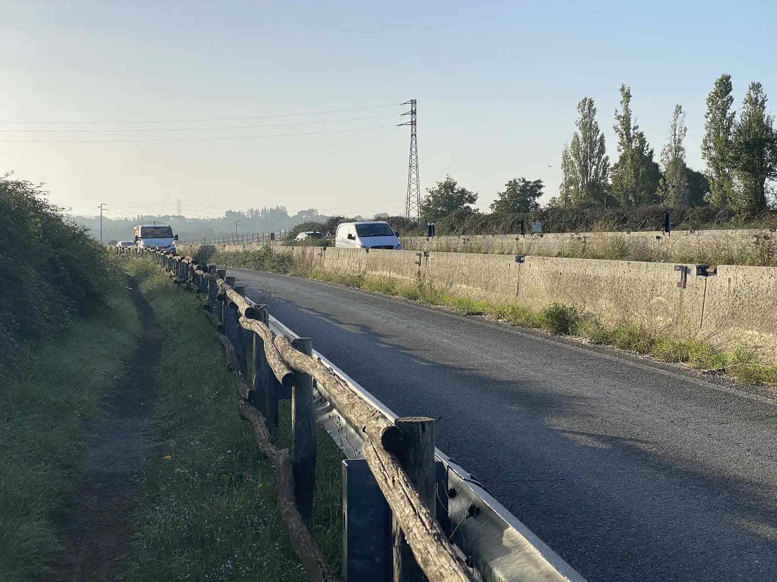 A footpath runs alongside a wooden guardrail beside a paved road, with white vans parked and a pylon on the horizon.