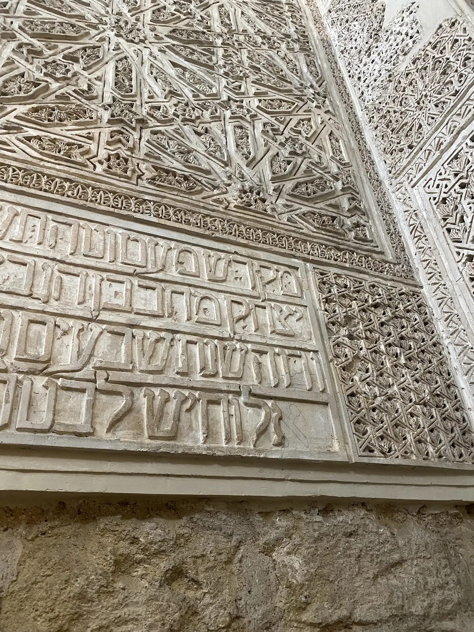 Carved plasterwork bearing a band of Hebrew inscription beneath panels of interlaced geometric and floral tracery, above bare stone walling.