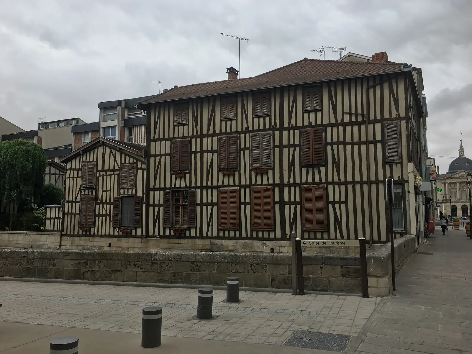 A row of half-timbered black-and-cream buildings on a street corner, a sign pointing left reading 'Office de Tourisme' and a domed civic building visible further down the street.