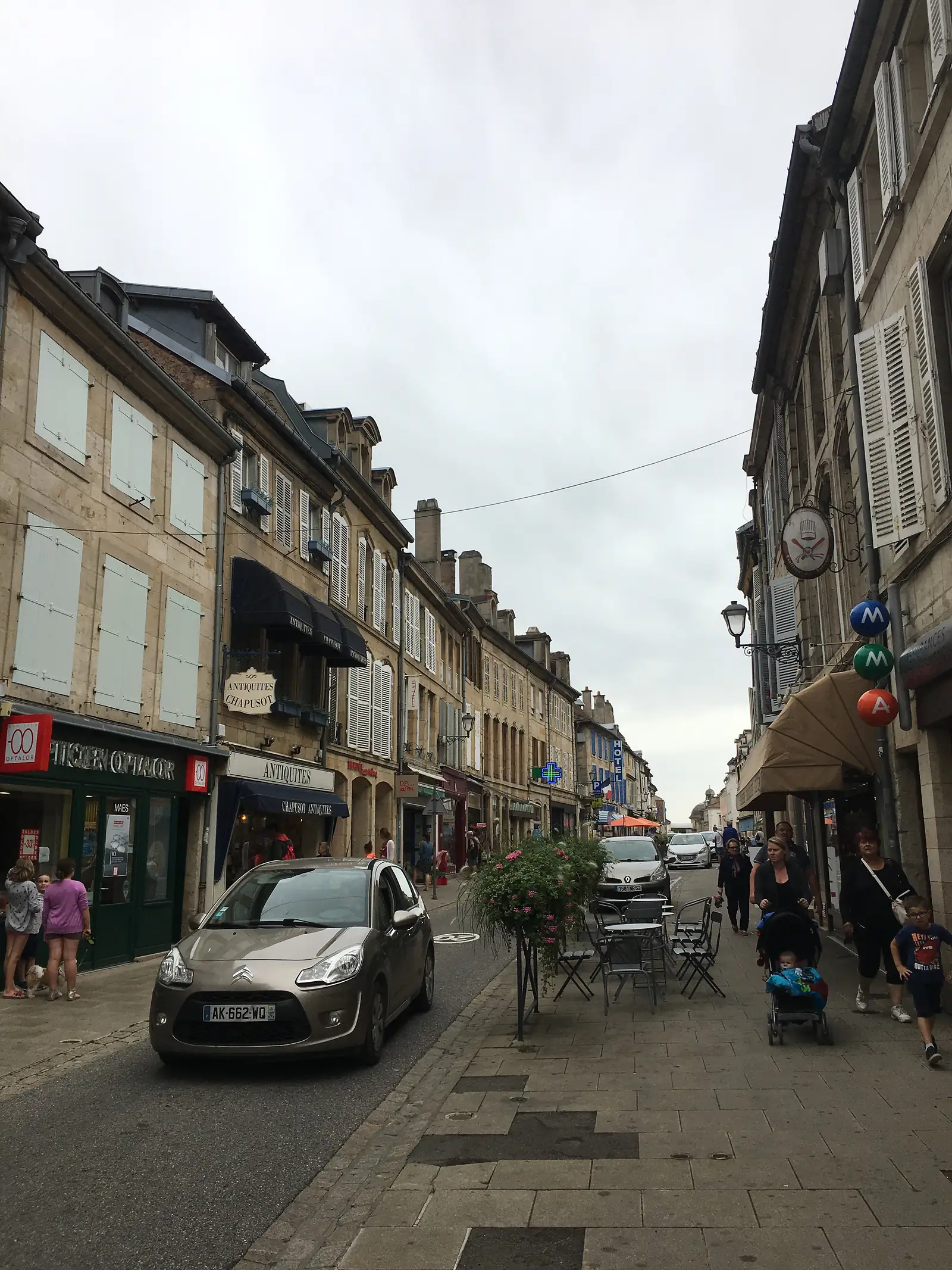 A pedestrianised shopping street in Langres lined with pale stone buildings, shop awnings and a hotel sign, with pavement café tables and shoppers strolling past.
