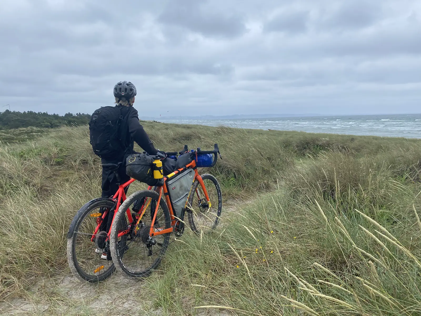 A cyclist with a rucksack standing beside two loaded bikes on a sandy dune path, looking out at a windy grey sea.