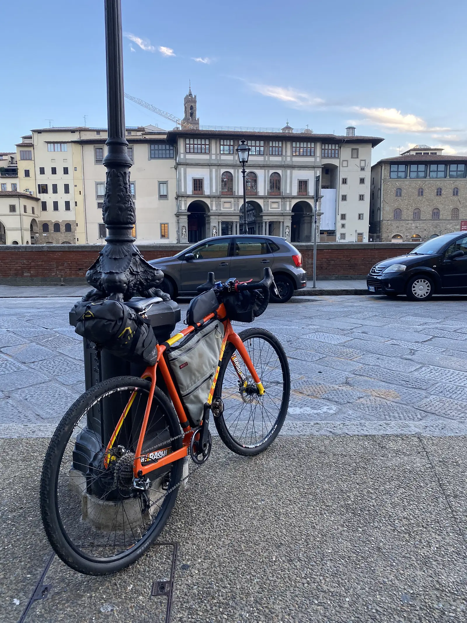 A loaded orange bikepacking bike leaning against an ornate lamppost base on a cobbled city street, with a church tower visible among the buildings beyond.