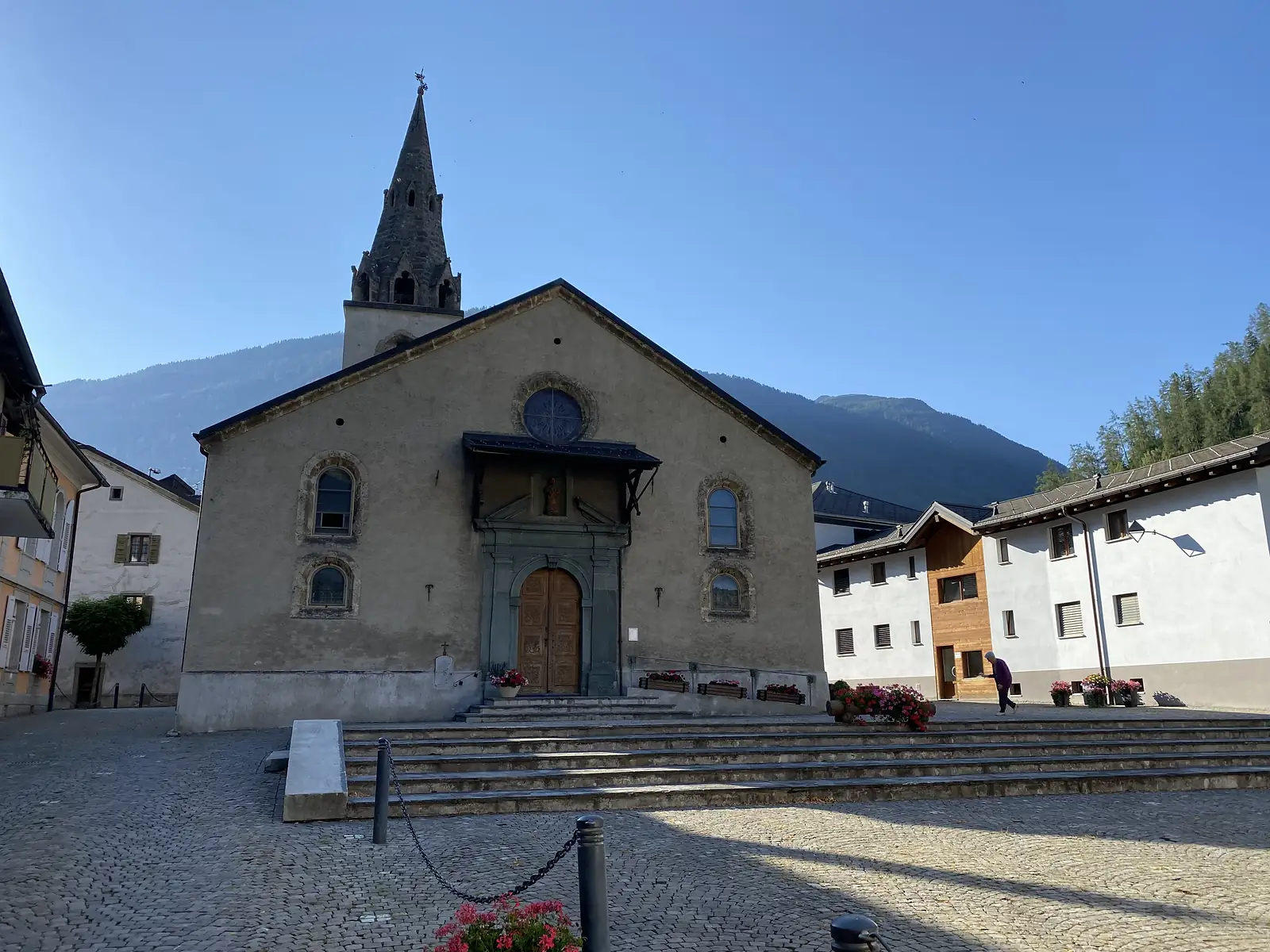 The stone facade and pointed bell tower of a village church above a cobbled square, with a person walking past flower-filled planters in early morning light.