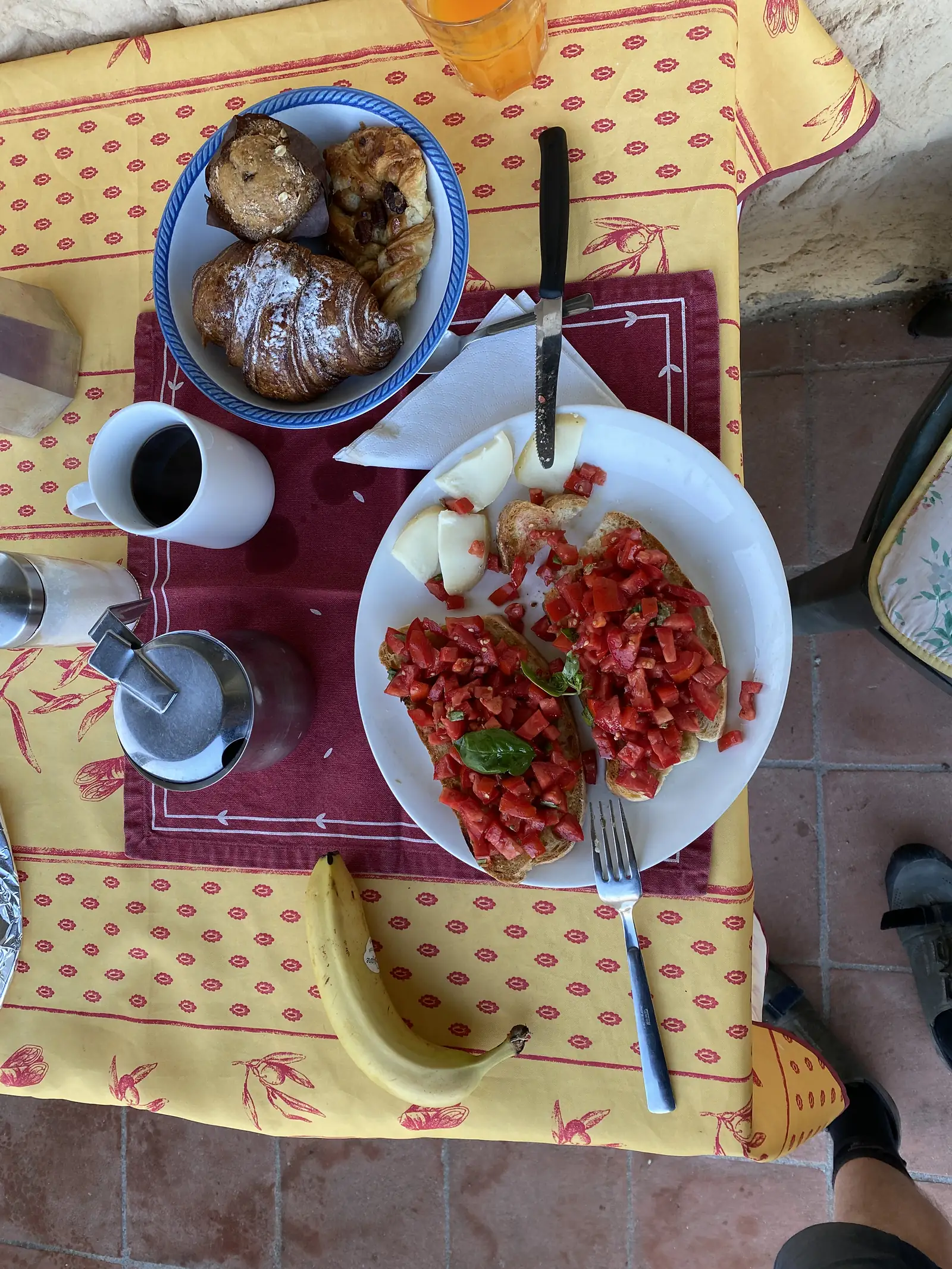 Overhead view of a breakfast table with tomato and basil bruschetta on toast, wedges of cheese, a bowl of pastries, a banana, orange juice and coffee, on a yellow patterned tablecloth.