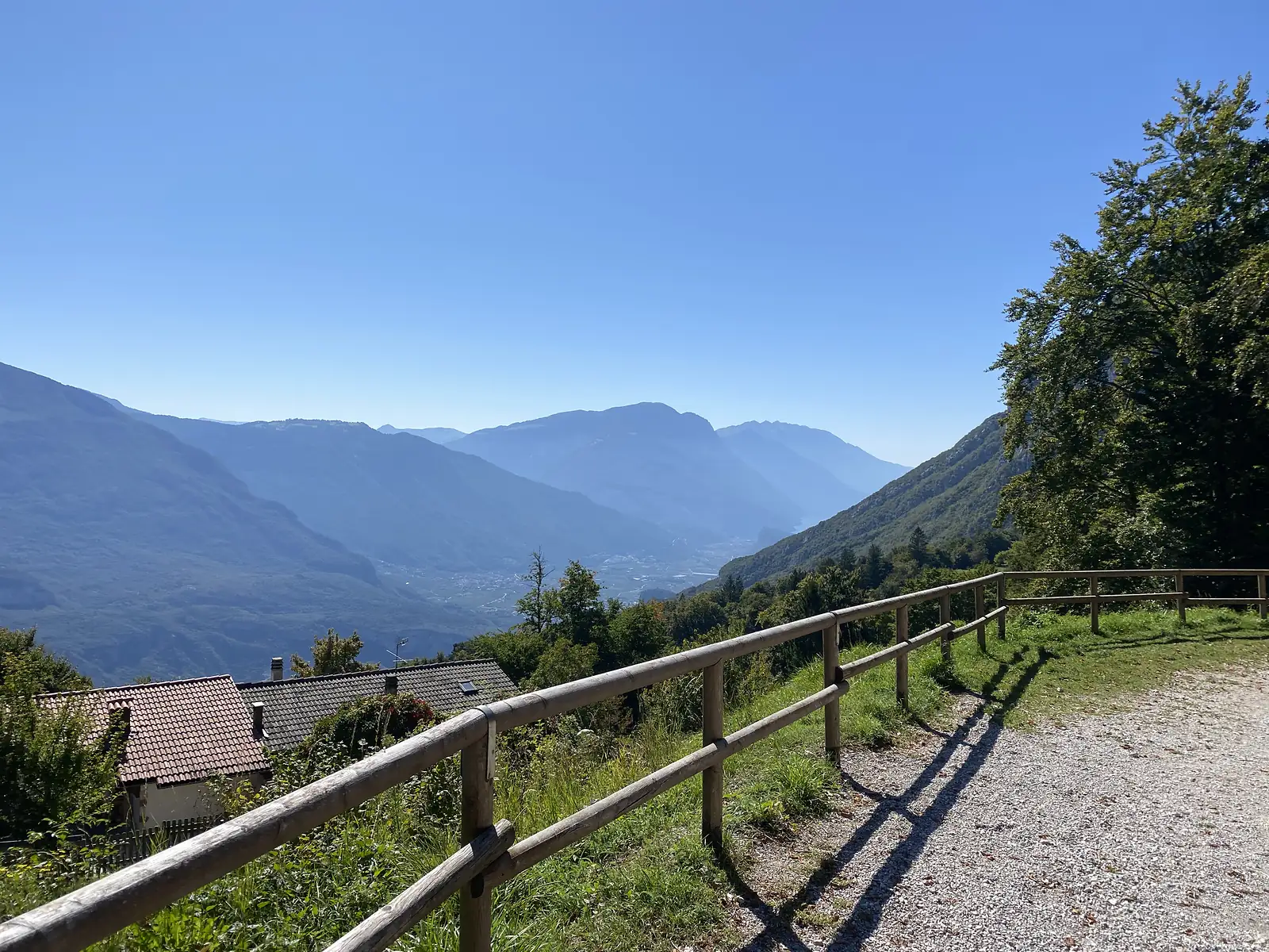A gravel path behind a timber fence looks out over tiled rooftops to a wide hazy valley between mountain ridges.