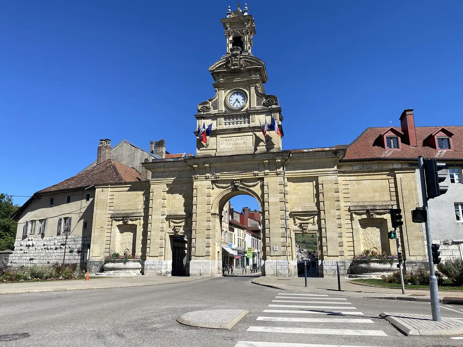 A grand stone gateway topped with an ornate clock tower and inscribed 'REPUBLIQUE FRANÇAISE' spans a street lined with shuttered buildings, likely marking the approach to Pontarlier at journey's end.