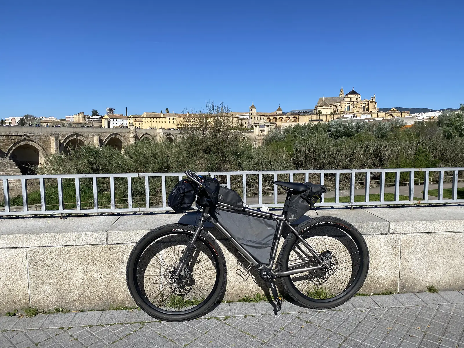 A loaded bikepacking bike propped against a white railing, with an ancient many-arched stone bridge and a domed cathedral skyline behind.