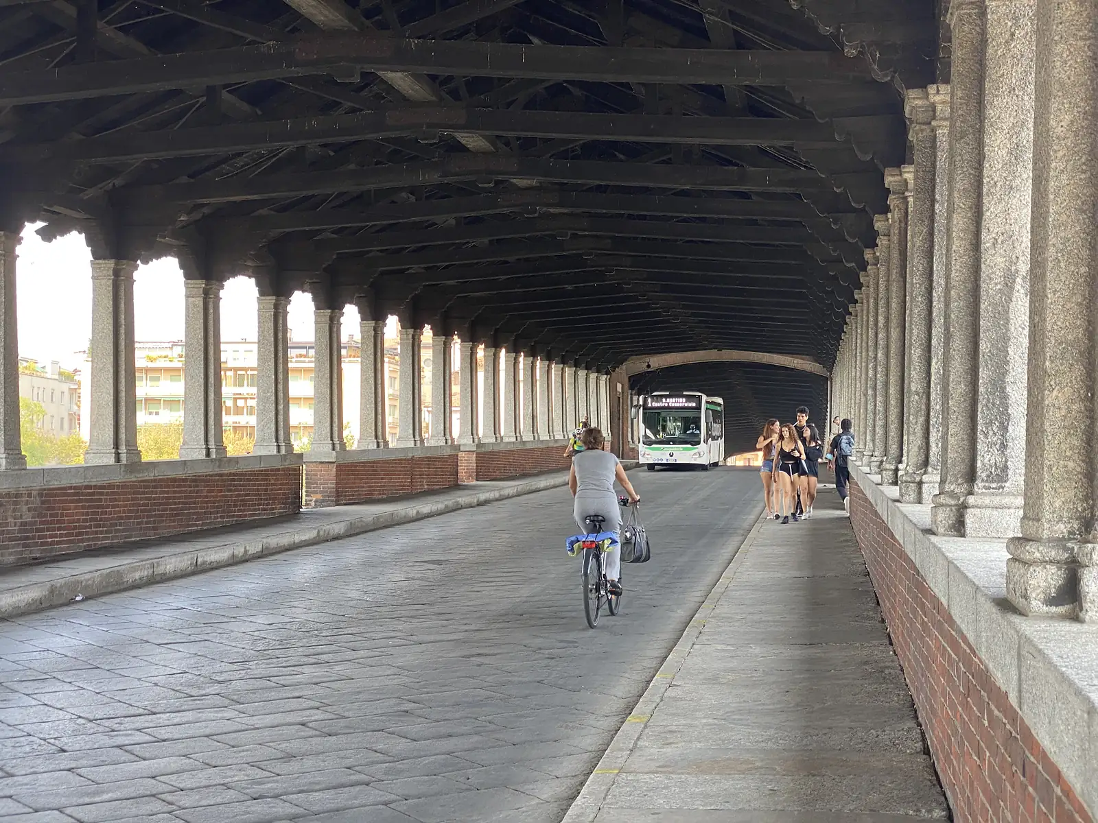 Cyclists and pedestrians share the covered walkway of Pavia's Ponte Coperto, its stone columns lining a road along which a bus approaches.