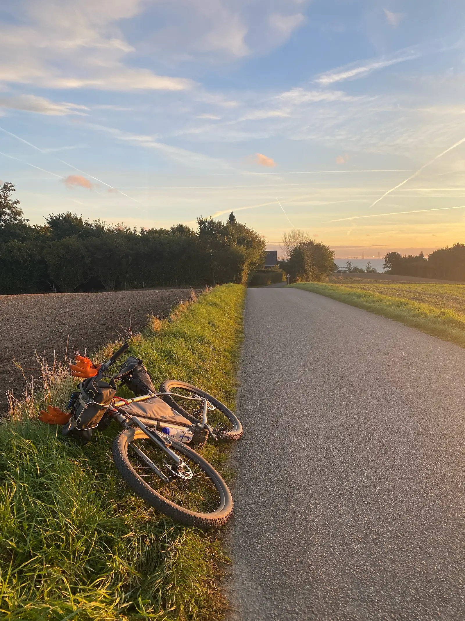 A loaded bikepacking bike laid on the grass verge of an empty country road at sunset, with hedgerows and the sea beyond.