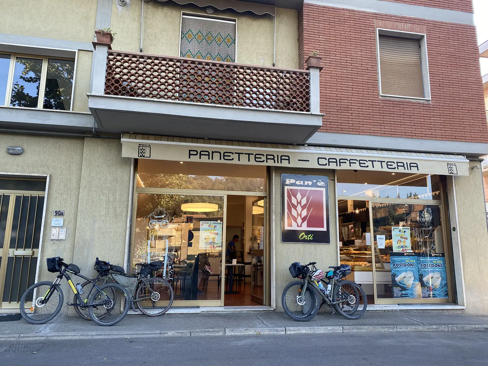 Four loaded touring bikes are parked outside a bakery and café with a striped awning reading "PANETTERIA — CAFFETTERIA" and a "Pan'è" sign.