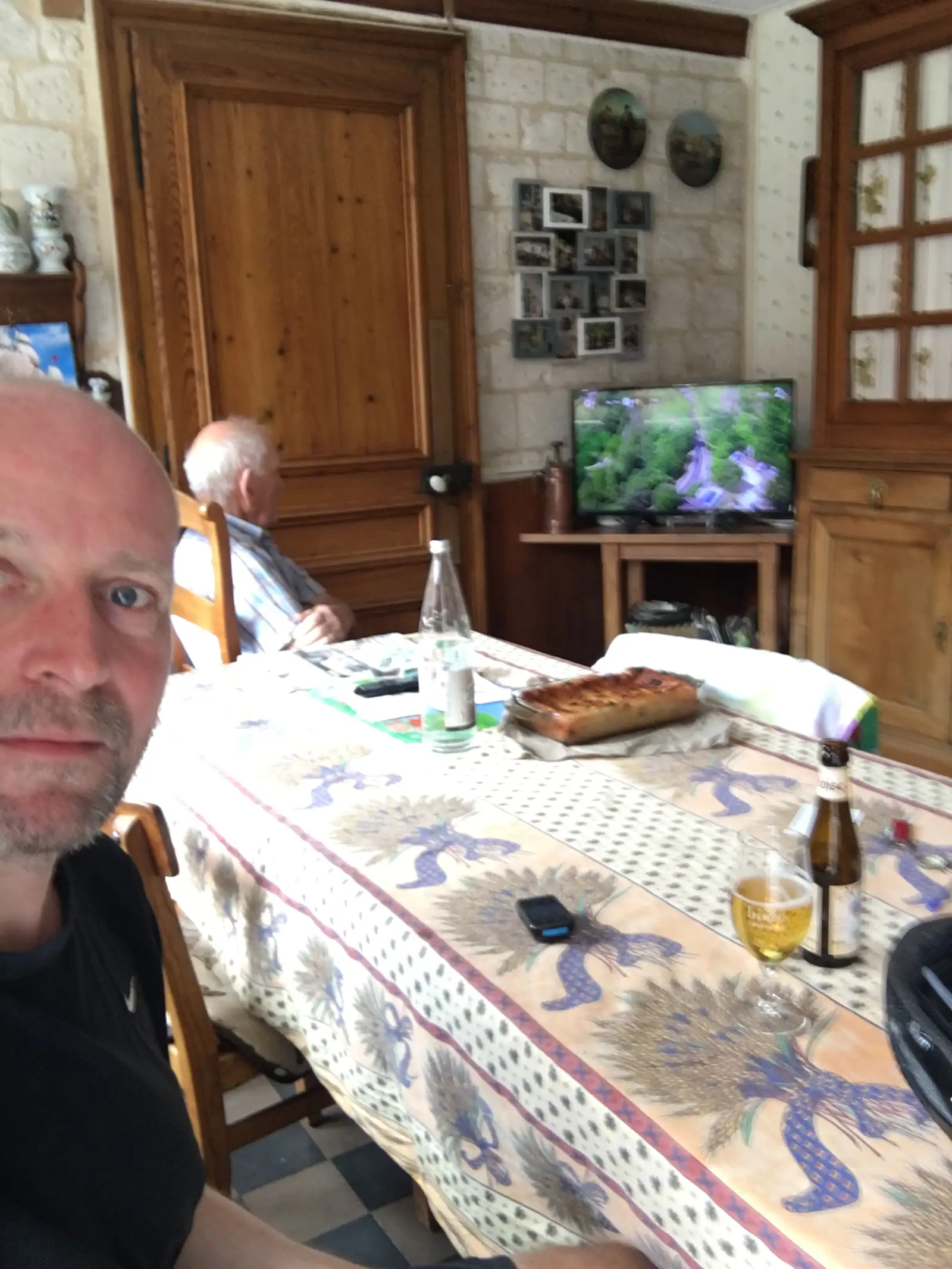 A cyclist takes a selfie at a dining table set with beer, a slice of tart and a glass, with an older man seated behind him in a kitchen with a television and family photographs.