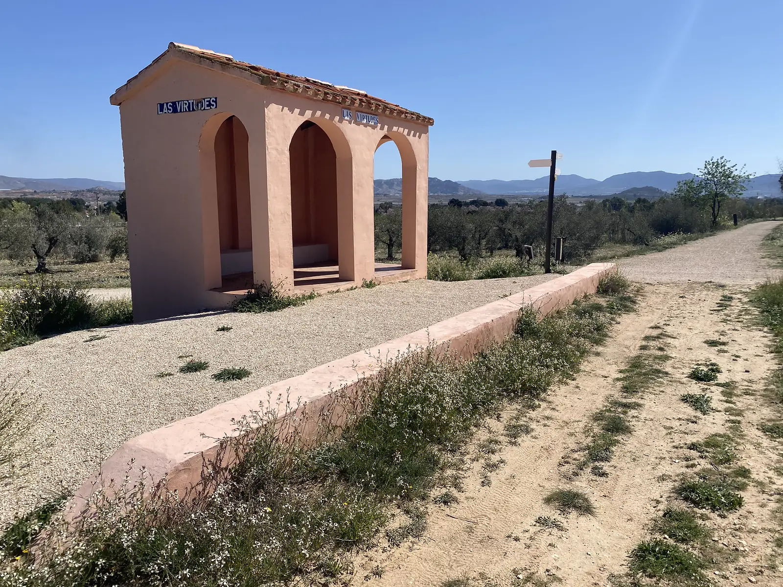 A pink-rendered shelter of three arched bays under a tiled roof, its tiled plaques reading "LAS VIRTUDES", beside a gravel path through an olive grove.