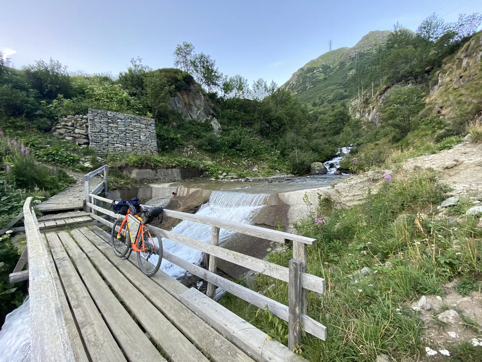 A loaded touring bike stands on a wooden footbridge beside a small weir where a mountain stream tumbles over concrete steps.