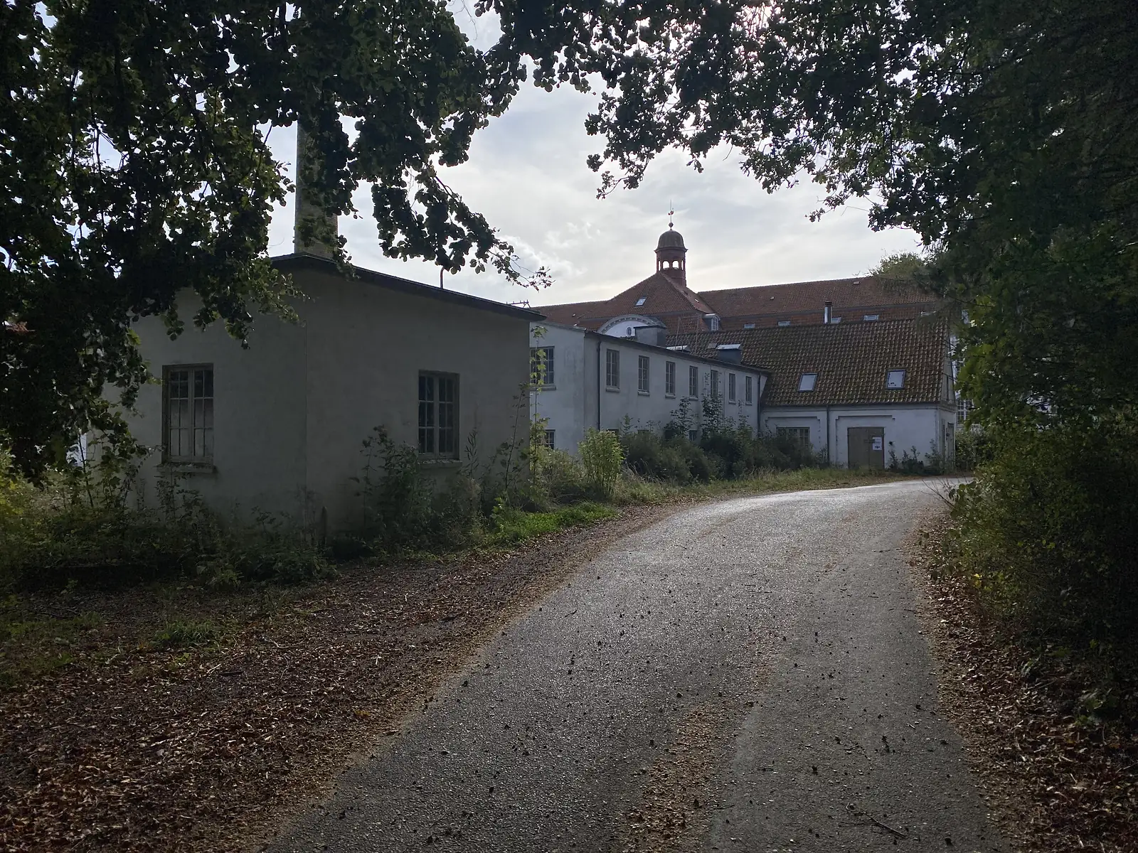 A gravel drive curving past a disused rendered outbuilding with a tall chimney towards a long white manor range with a lantern turret.