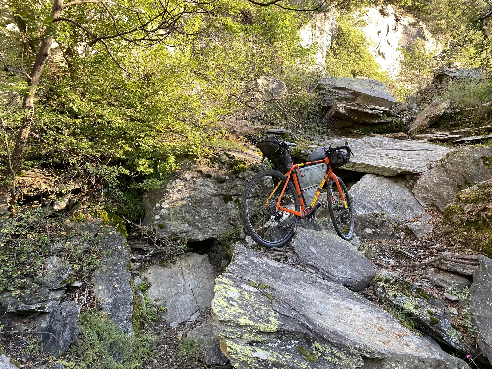 A loaded orange gravel bike propped upright among large flat boulders on a steep, rocky hillside track.