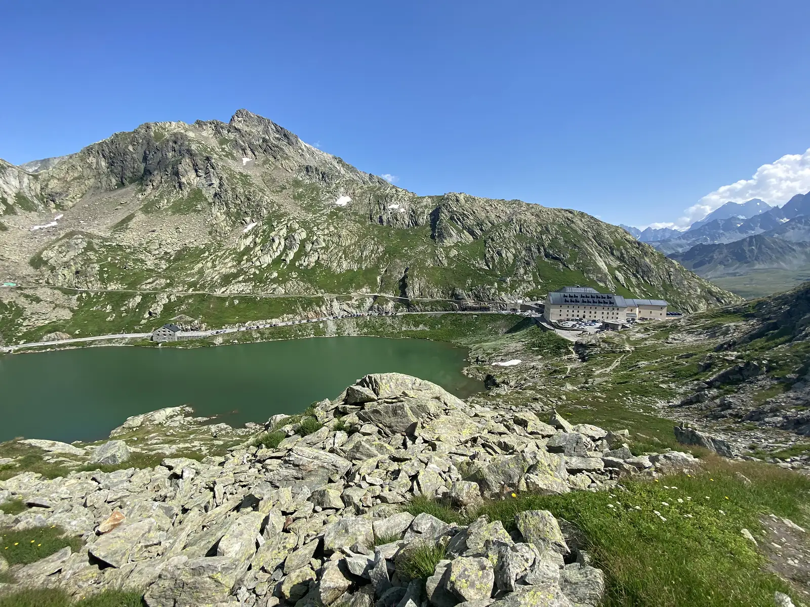 A rocky outcrop overlooking the green pass lake and hospice, with a single sharp peak rising behind the valley road.