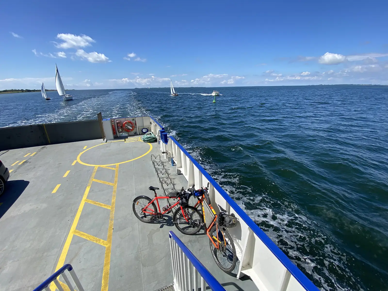 Two loaded bikes strapped to a rack on a ferry's open stern deck, the wake streaming away and yachts under sail on a blue sea.