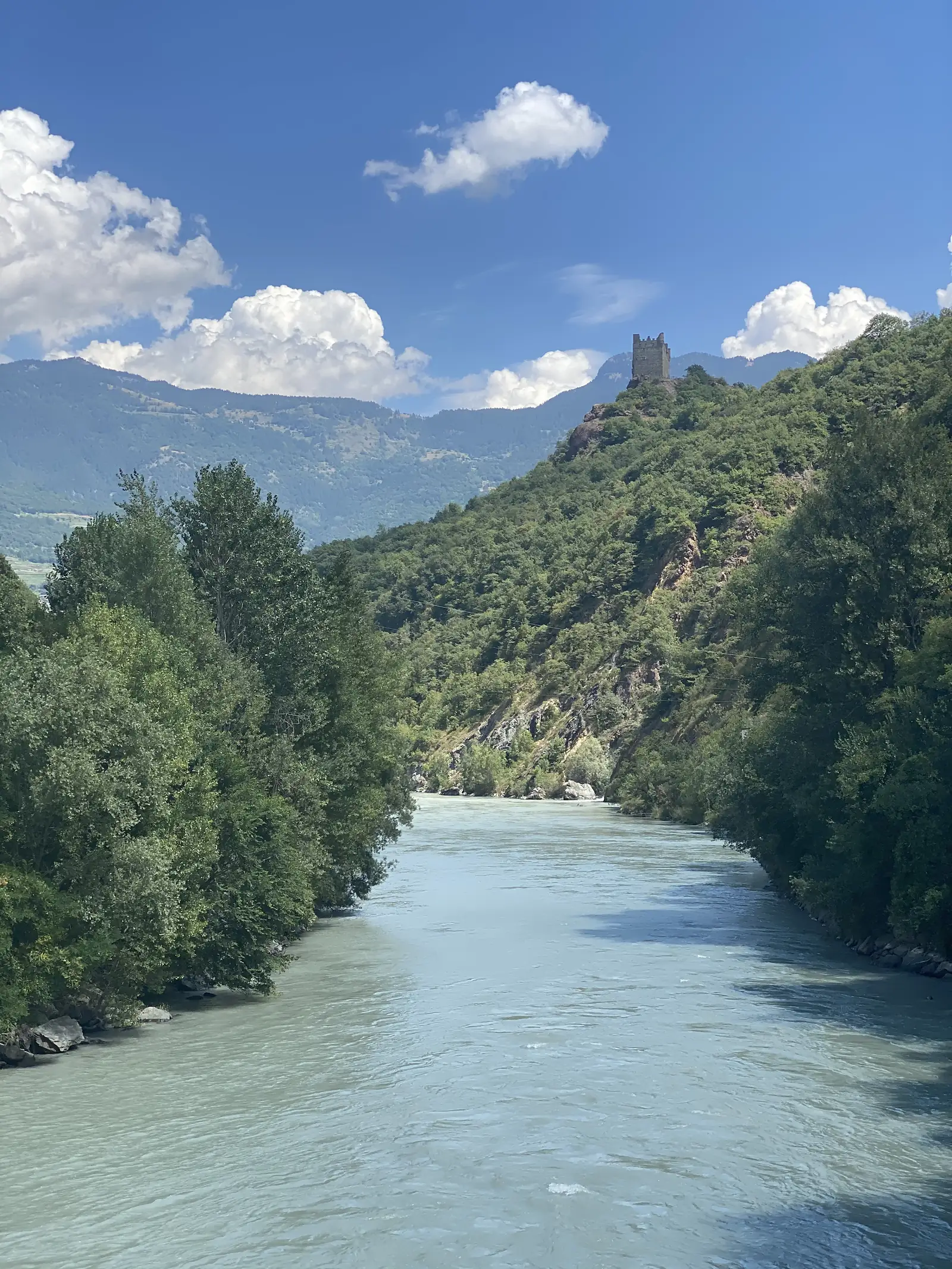 A pale turquoise river winds below a wooded ridge, a ruined square castle tower perched on a rocky outcrop above the trees near Saint-Vincent.
