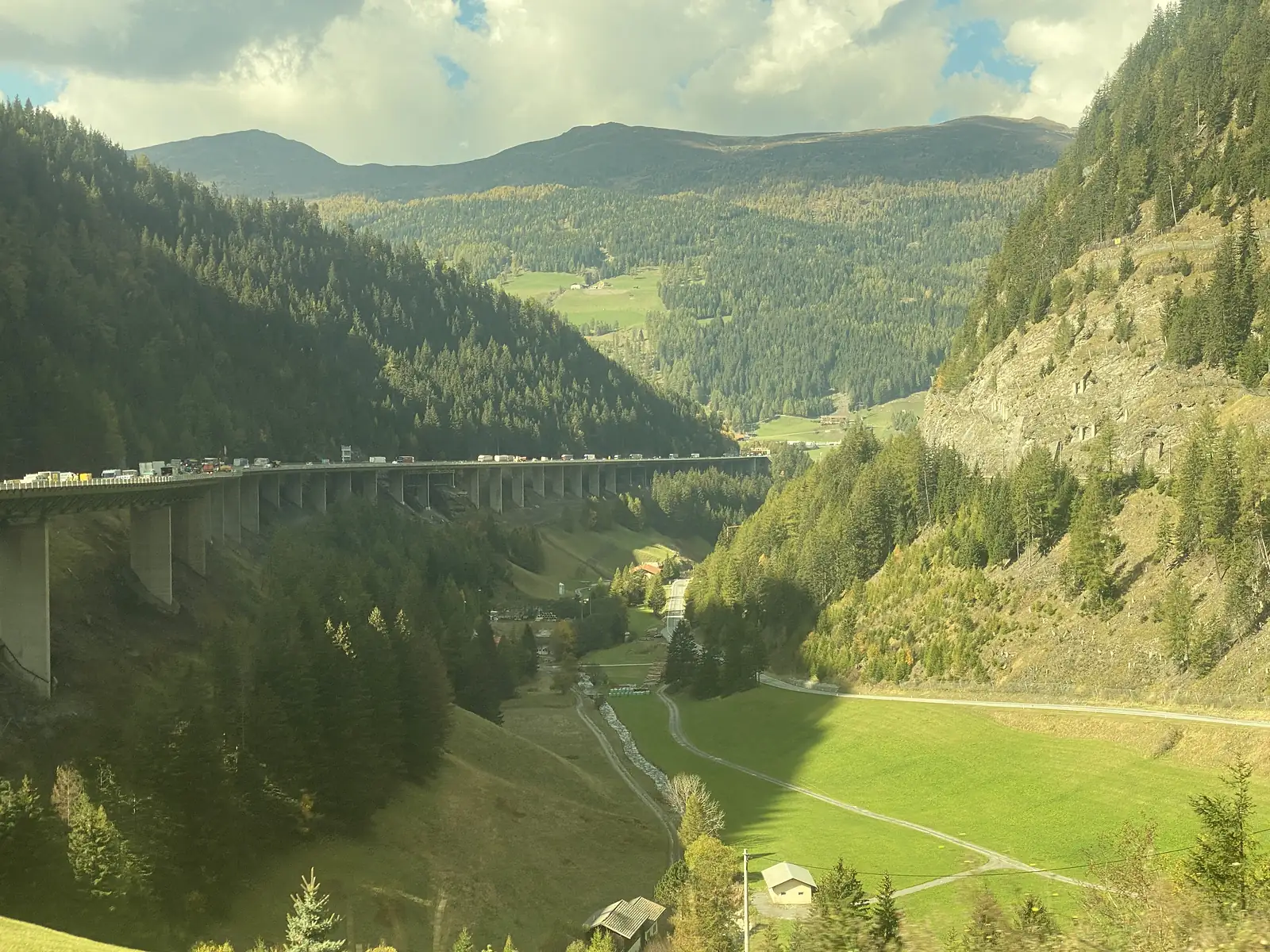 Heavy motorway traffic crosses a tall concrete viaduct spanning a steep, tree-covered alpine valley.