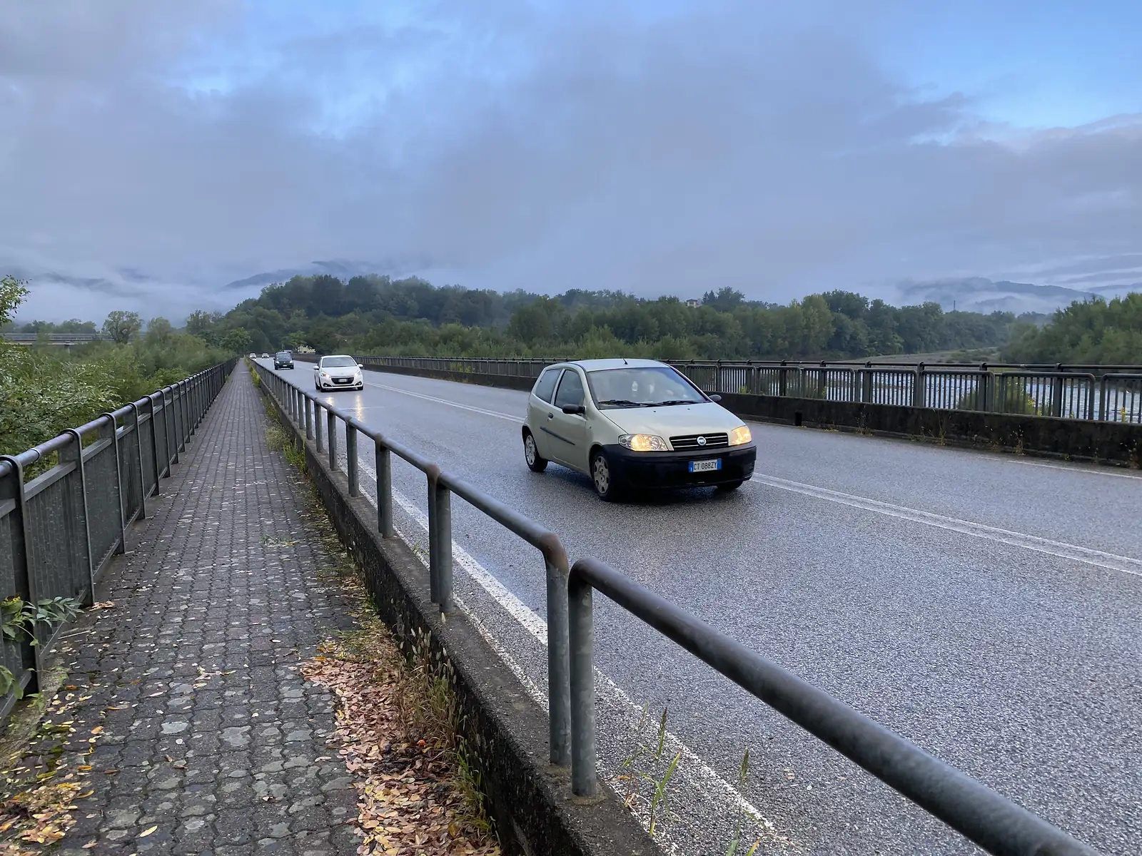 Cars cross a road bridge over a river running high and pale with silt, mist clinging to the hills beyond.