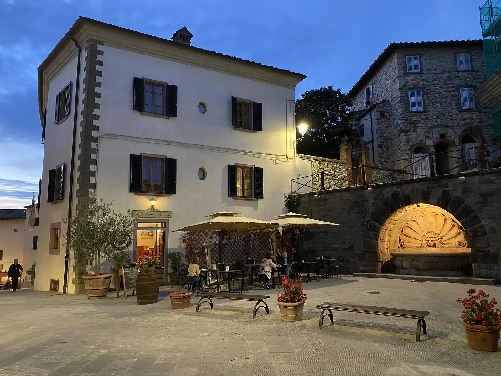 Diners seated under parasols outside Caffè San Niccolò at dusk, beside an ornate stone lion-head fountain set into an arched wall.