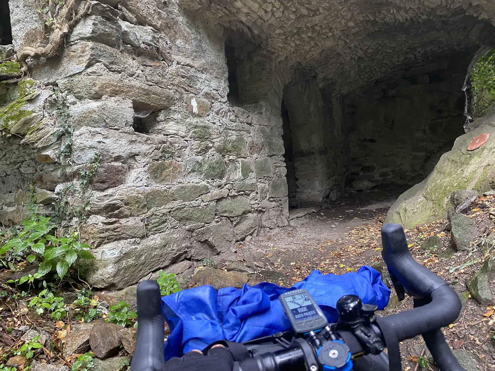 A rough stone archway and ruined wall mark the entrance to an old fortified passage, with a white waymark and red trail blaze visible on the rock.