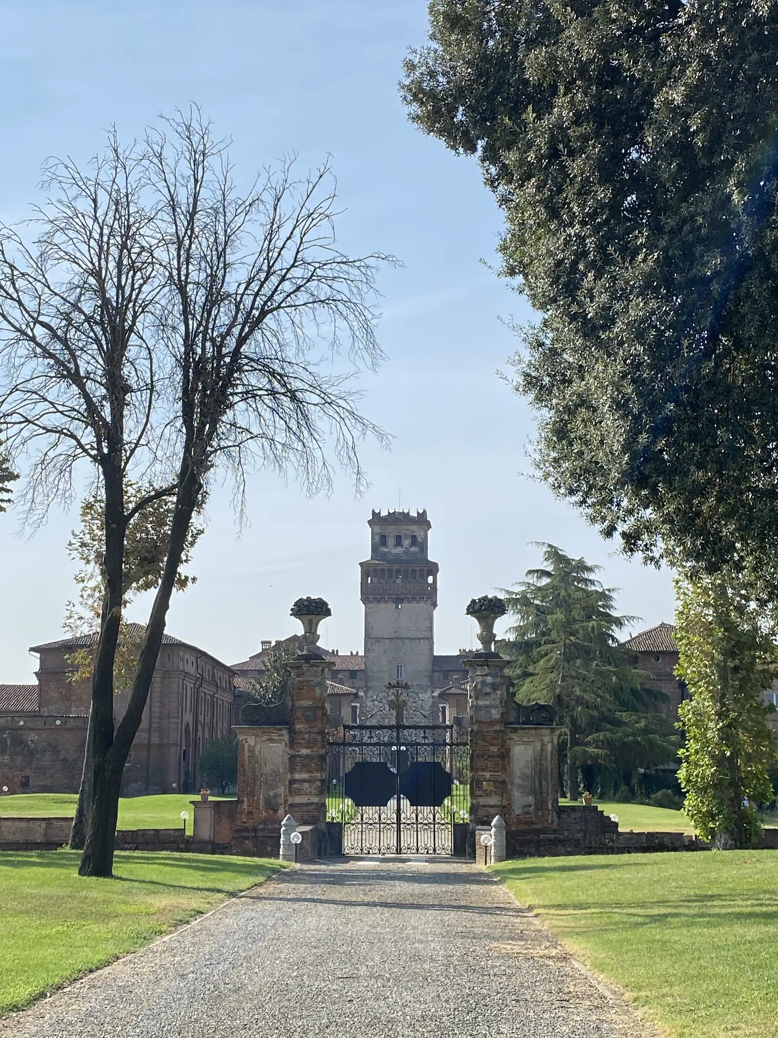 Stone gate piers topped with statues and urns flank the wrought-iron entrance to a grand villa, its crenellated tower rising behind trees.