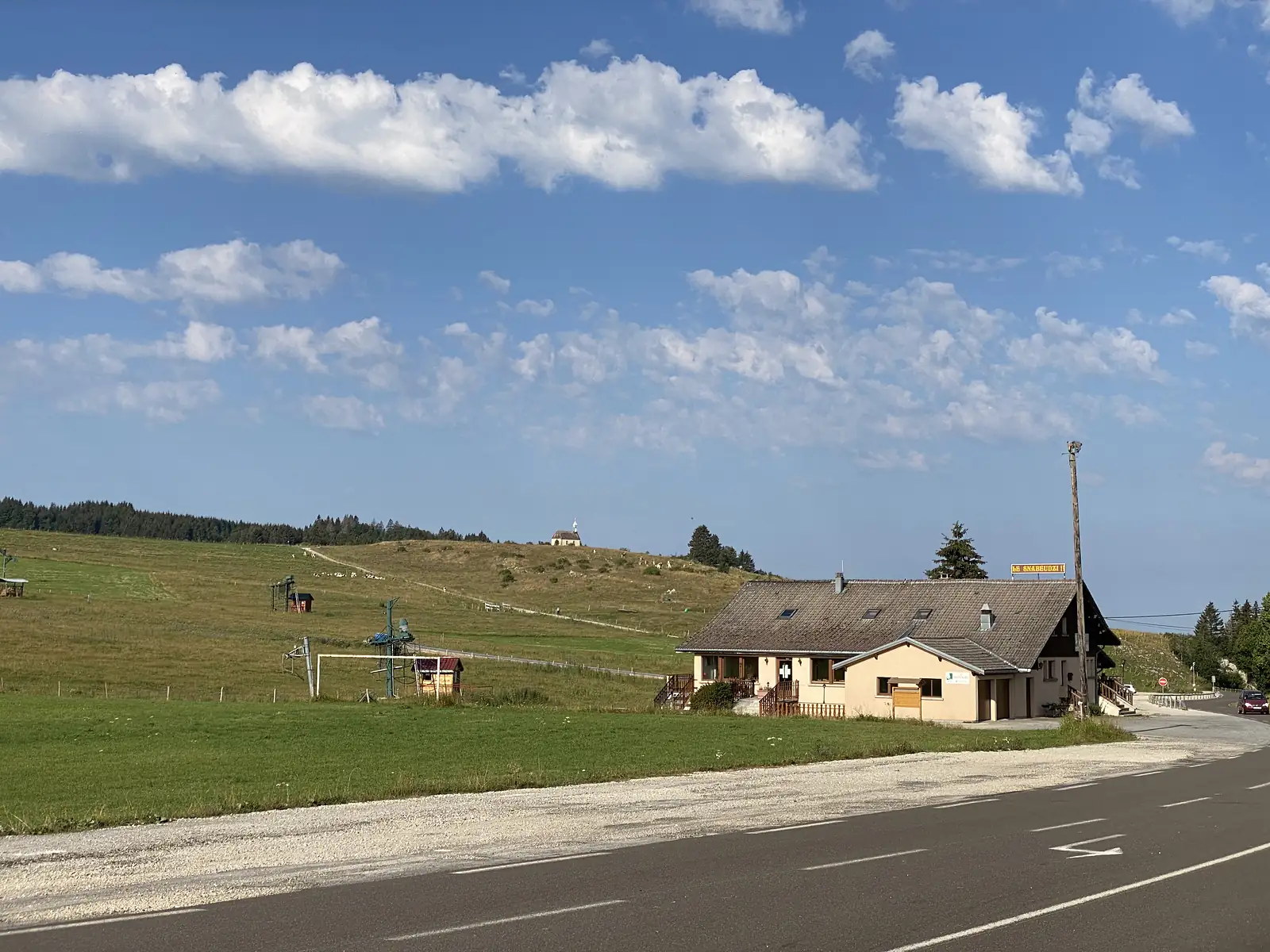 A hillside chalet-style building signed 'LB Snabeudzi' beside grazing pasture, with disused ski-tow pylons in the foreground and a small chapel on the ridge beyond.