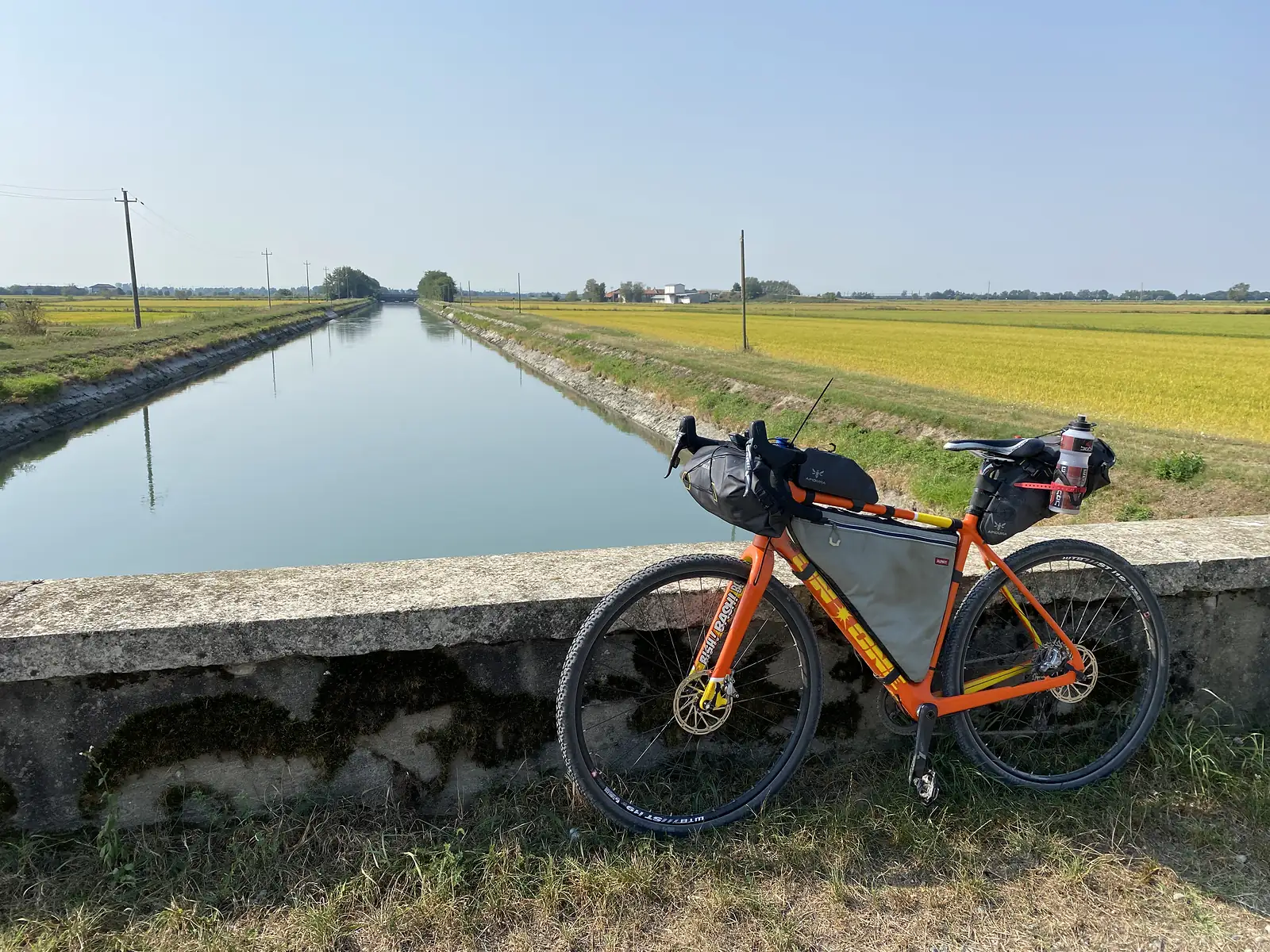 A loaded orange bikepacking bike propped on a low bridge parapet above a straight irrigation canal running between rice fields.