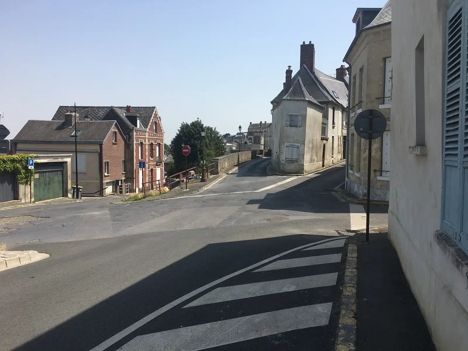 A quiet street of brick and stone gabled houses curves downhill past a no-entry sign, with open countryside visible in the distance.