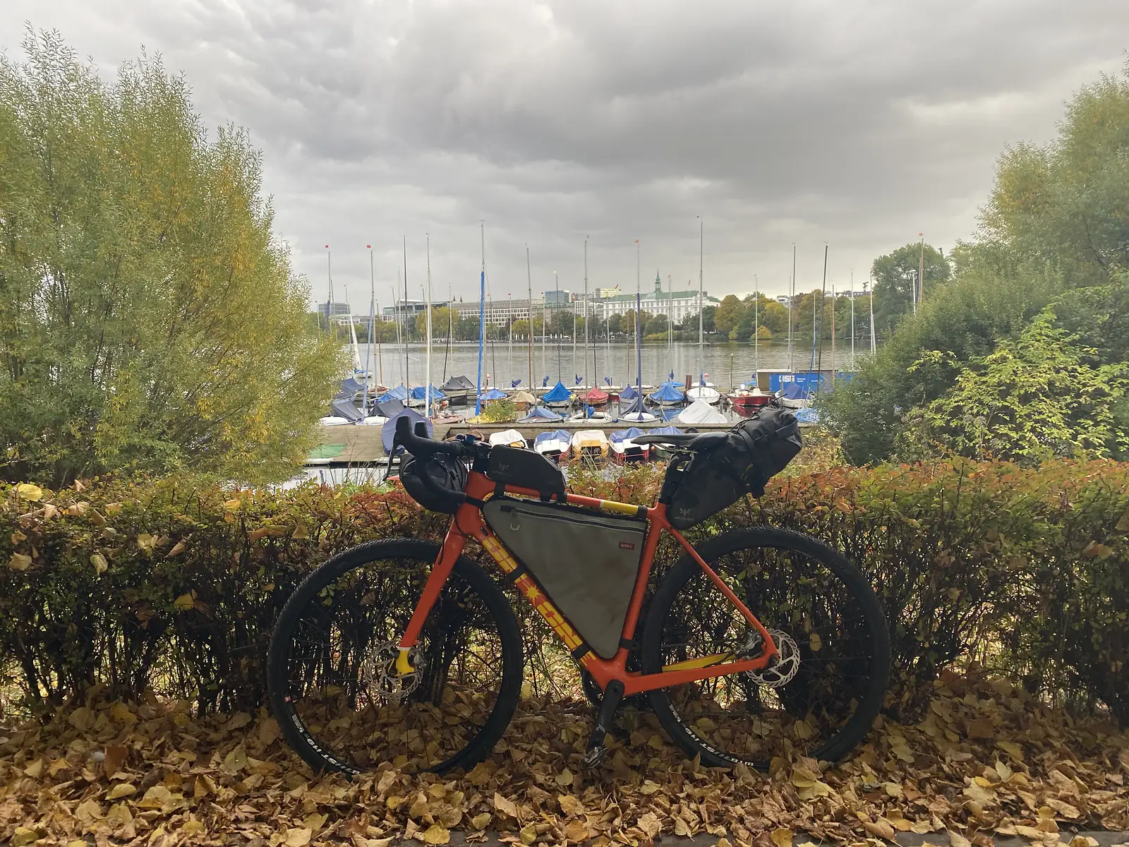 A loaded gravel bike stands against an autumn hedge overlooking a marina of moored sailing boats with a city skyline beyond.