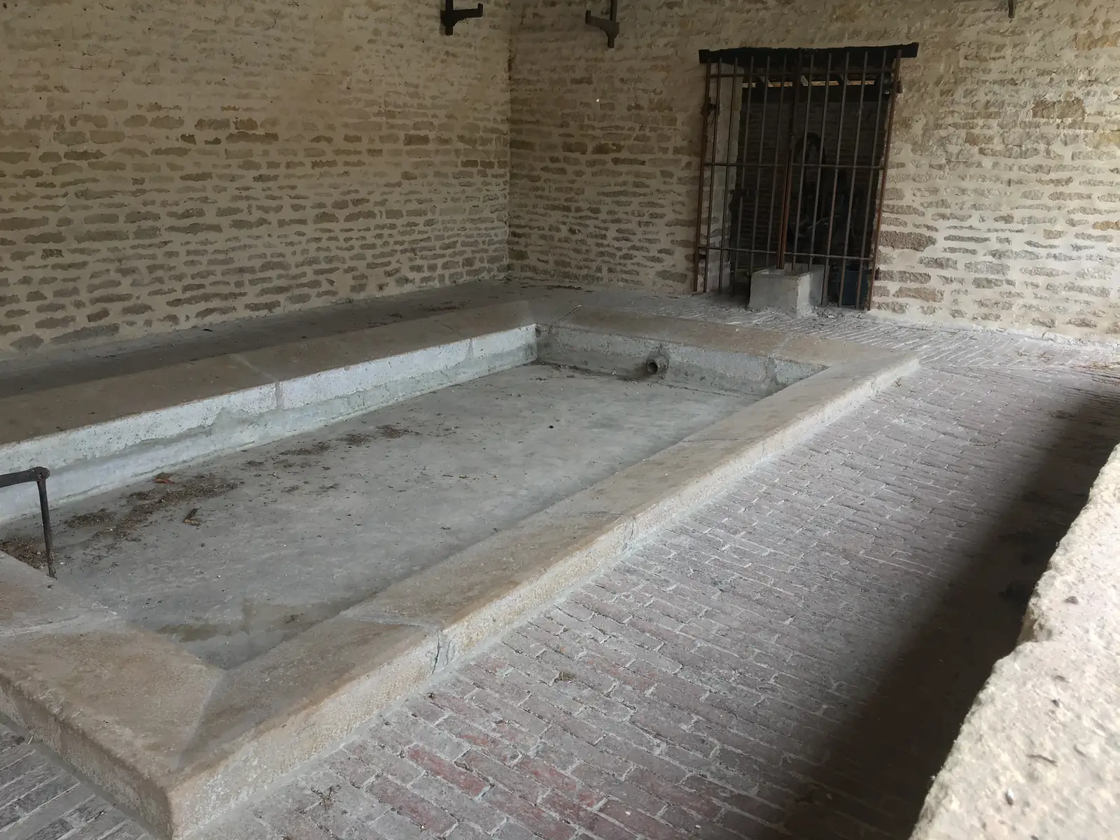 An empty stone-built wash house basin sits beneath a barred window inside a brick-vaulted lavoir.