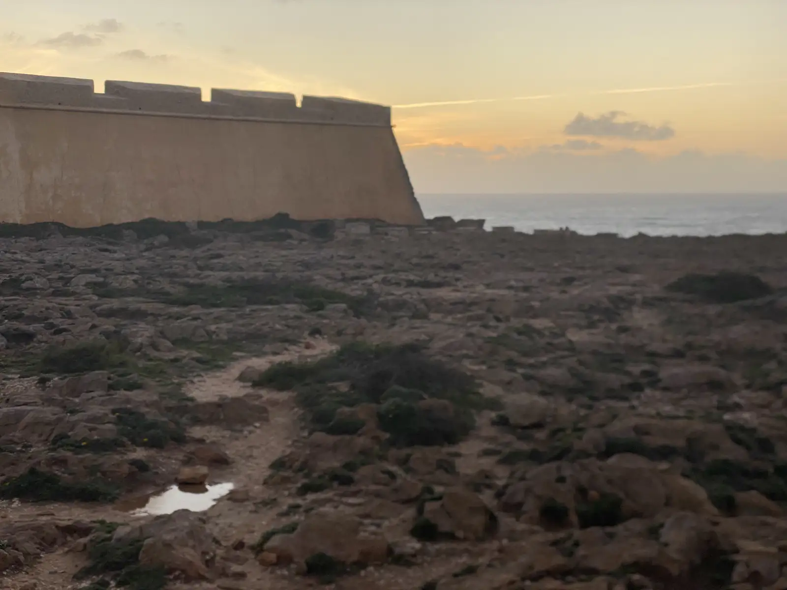 Sloping stone rampart of a clifftop fort catching the last light, with rough rocky ground and a rain puddle in the foreground and the sea beyond.