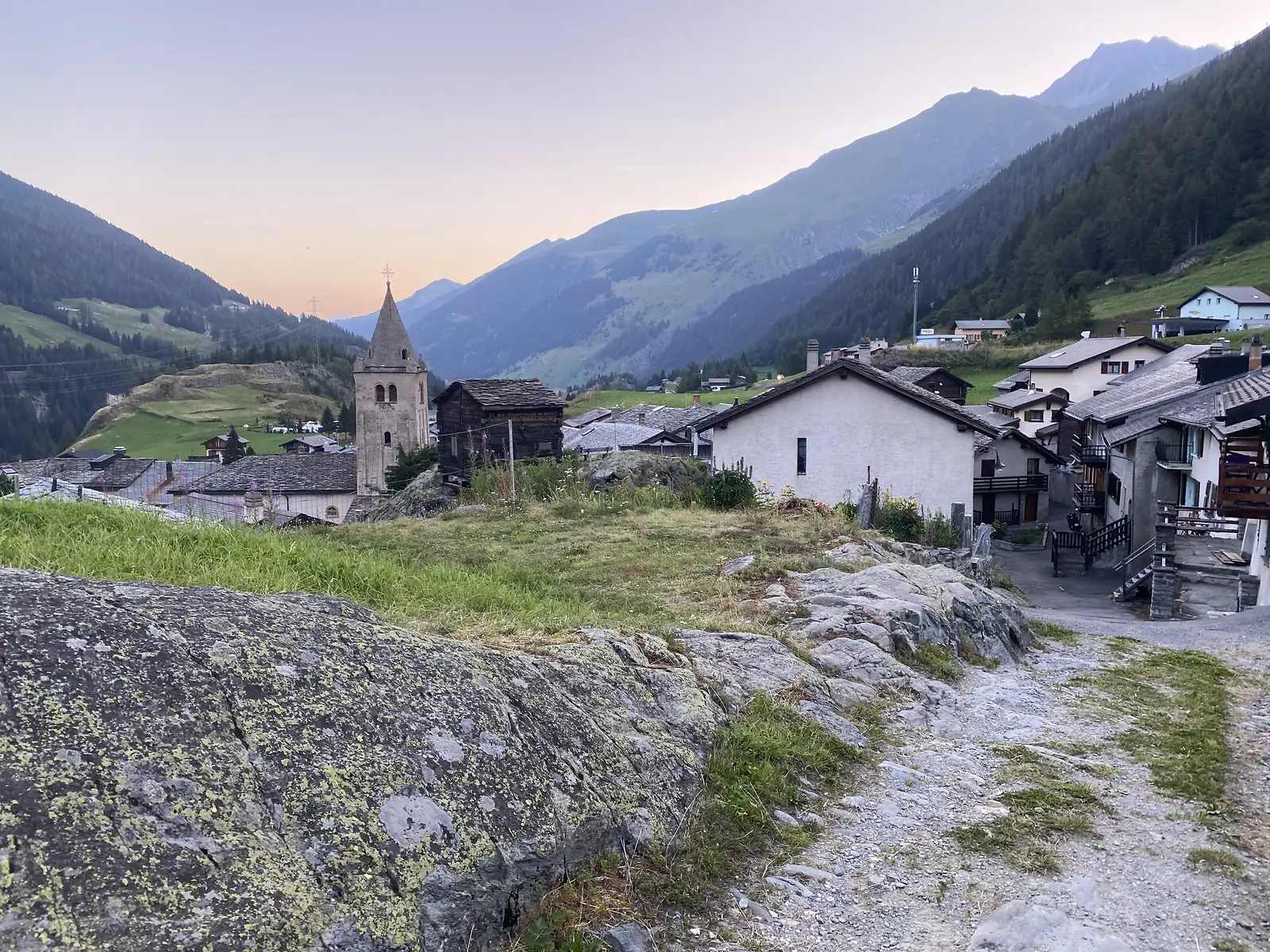 A stone church tower and cluster of stone-and-timber houses catch the last light at dusk, a mountain valley receding behind the Swiss village.