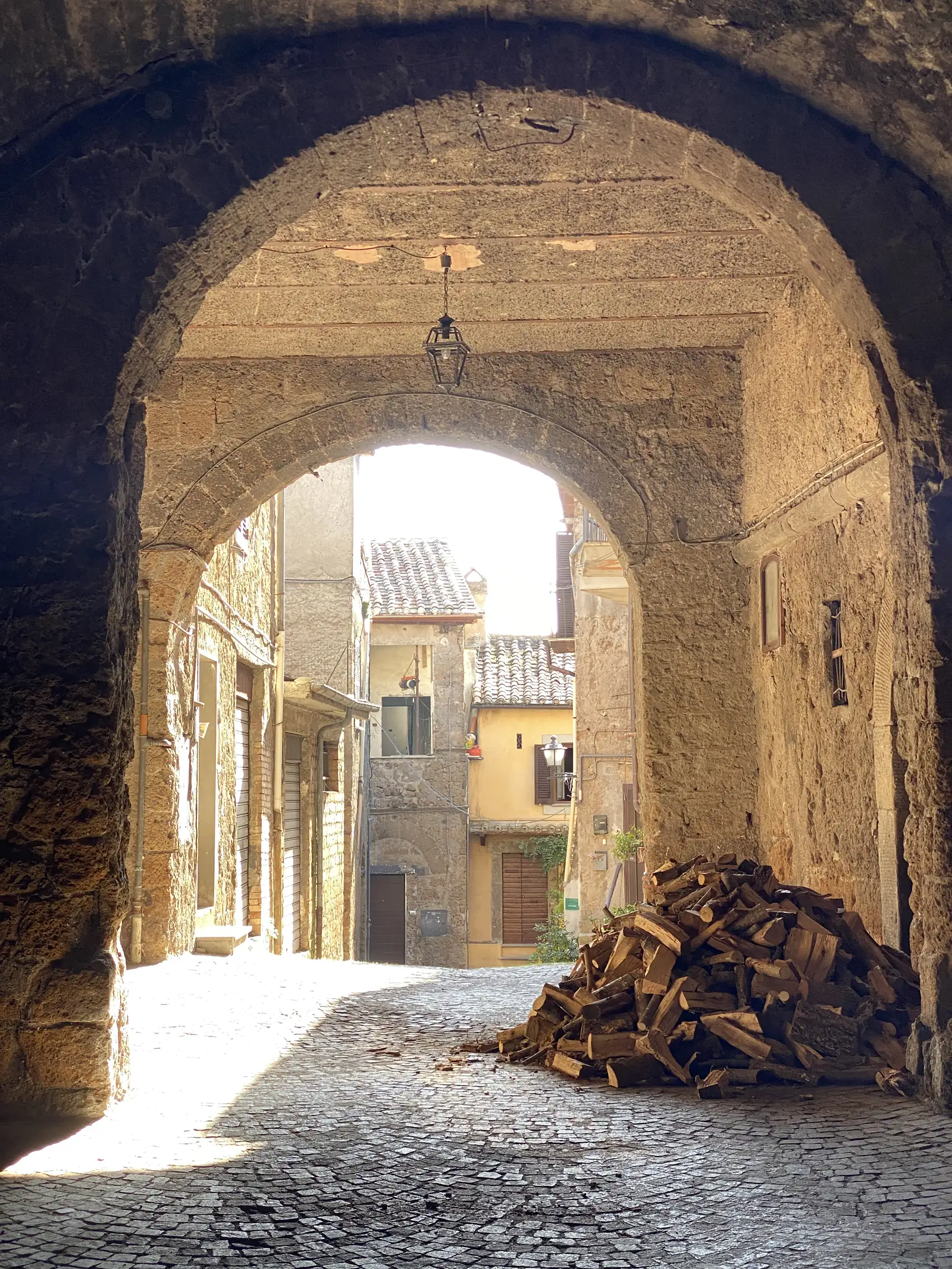 A vaulted stone passage through a medieval building opens onto a sunlit alley of stacked firewood and old townhouses beyond.