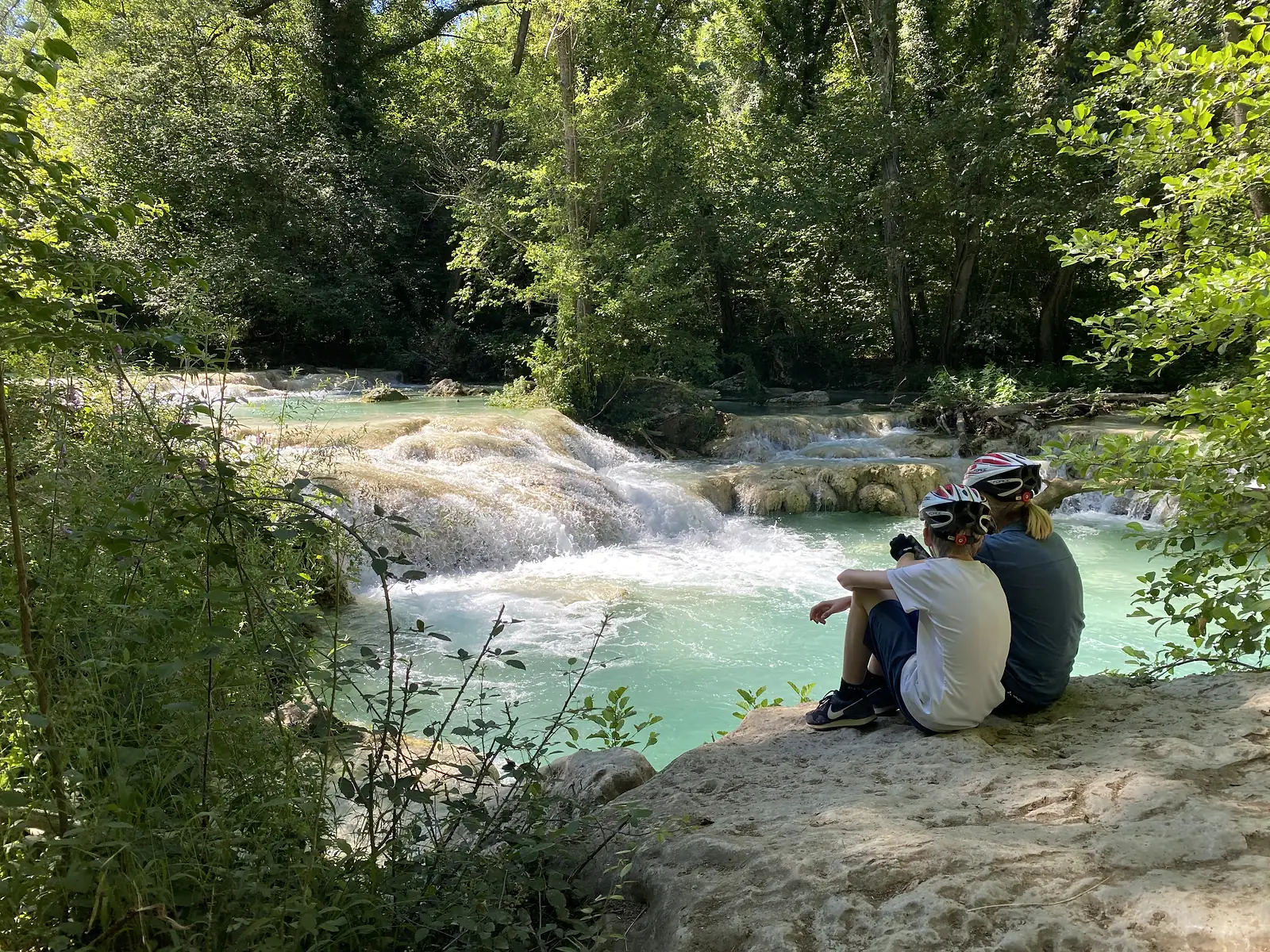 Two young cyclists in helmets sit on a rock ledge watching turquoise water tumble over travertine cascades in a wooded gorge.