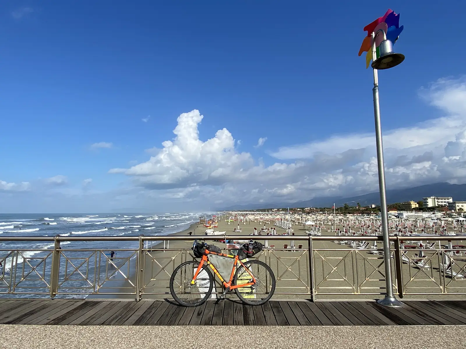A loaded orange bikepacking bike leans against a pier railing overlooking a beach crowded with umbrellas and sunbeds, a rainbow pinwheel spinning on a nearby lamppost.