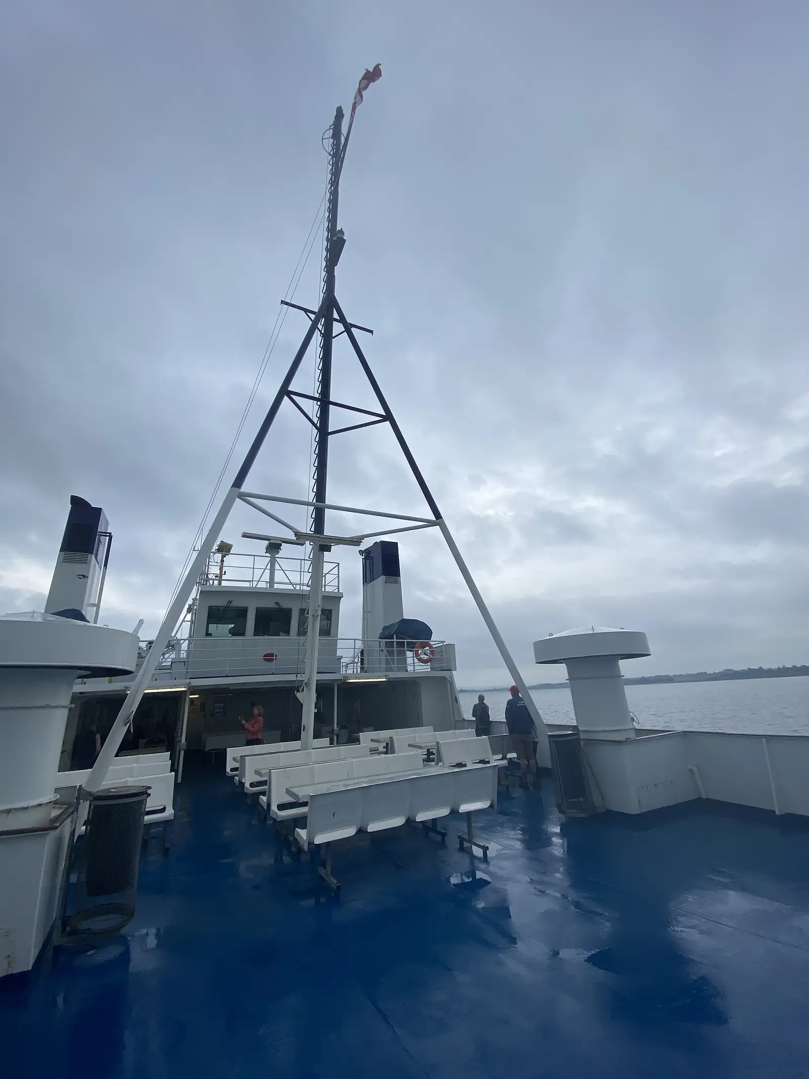 A ferry's open upper deck with a wet blue floor, white benches and a lattice mast, passengers standing at the rail under heavy cloud.