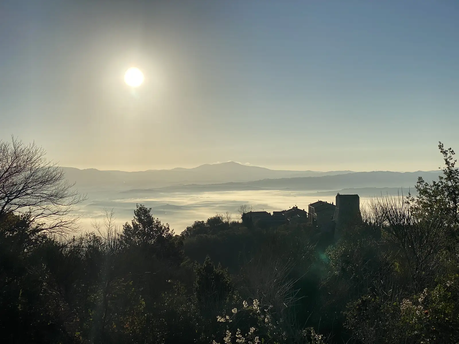 The sun rising above a hilltop hamlet whose buildings and tower stand in silhouette over a valley filled with morning mist.