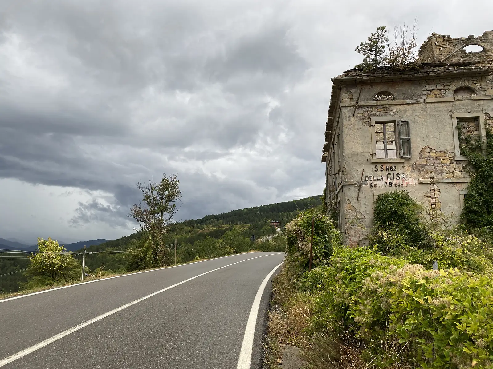 A crumbling abandoned building bears a weathered stencilled marker reading 'S.S. N.62 Della Cisa Km 79.047', standing beside a road that winds off into stormy hills.