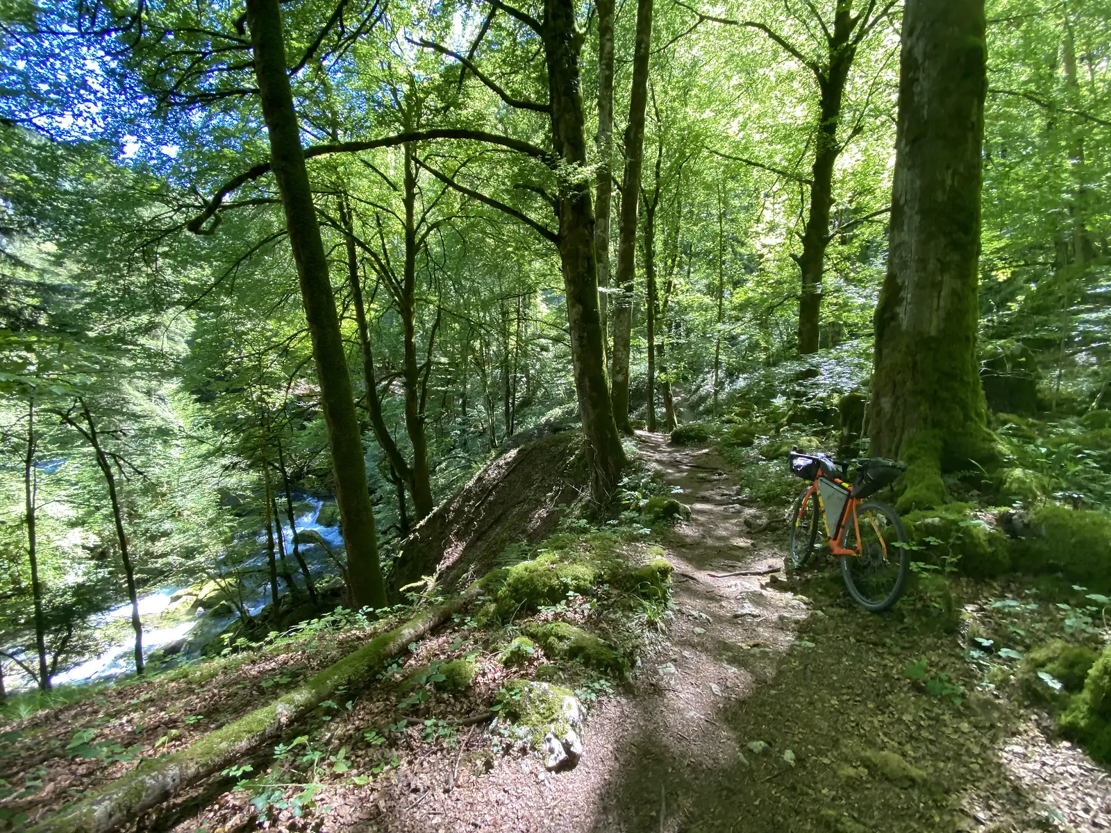 A loaded bike stands on a narrow woodland path cut into the slope above a turquoise stream tumbling over rocks in a gorge.
