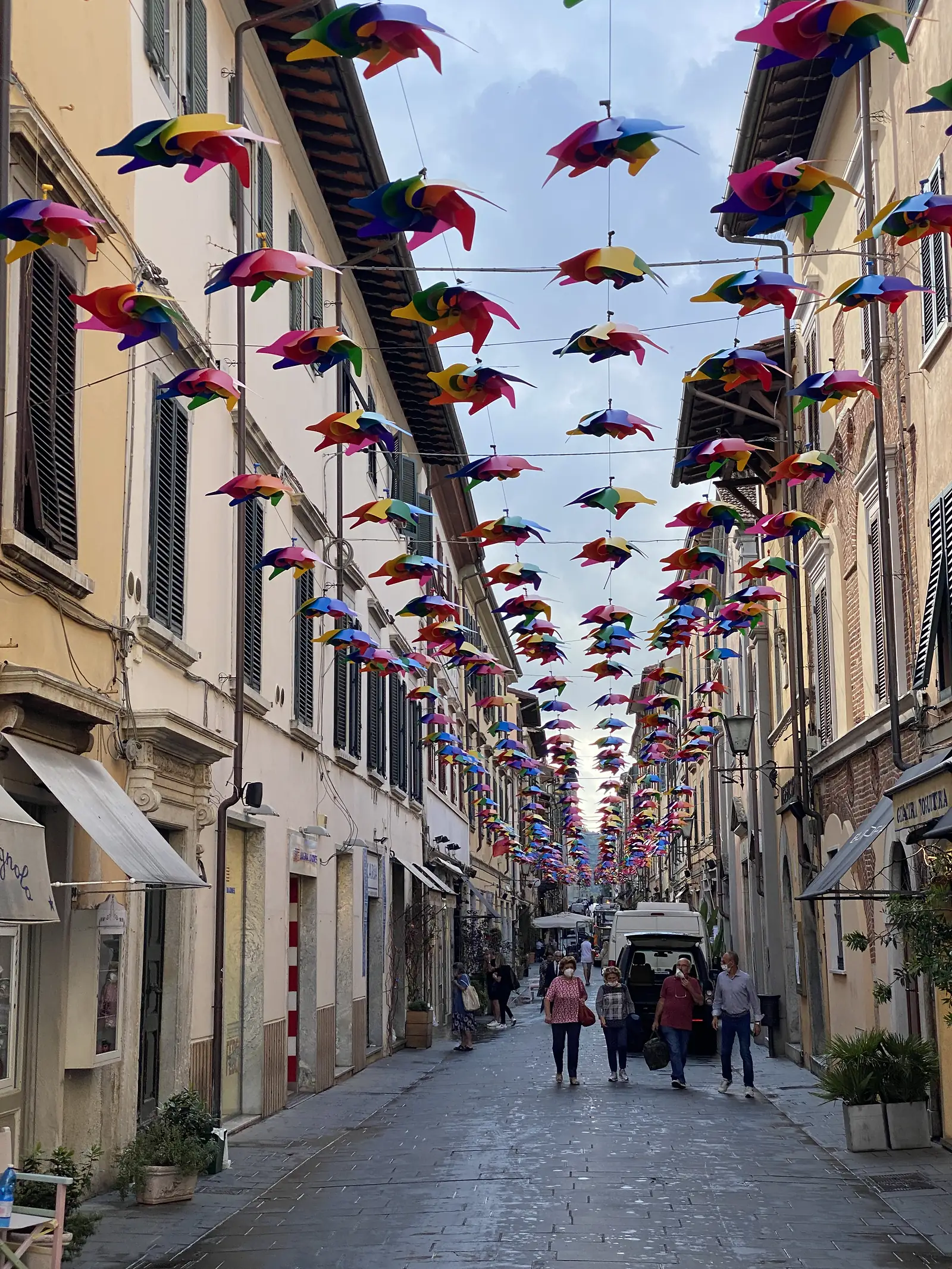A dense line of hanging pinwheels in rainbow colours stretches down a pedestrianised street between pastel-coloured buildings, with shoppers and a delivery van below.