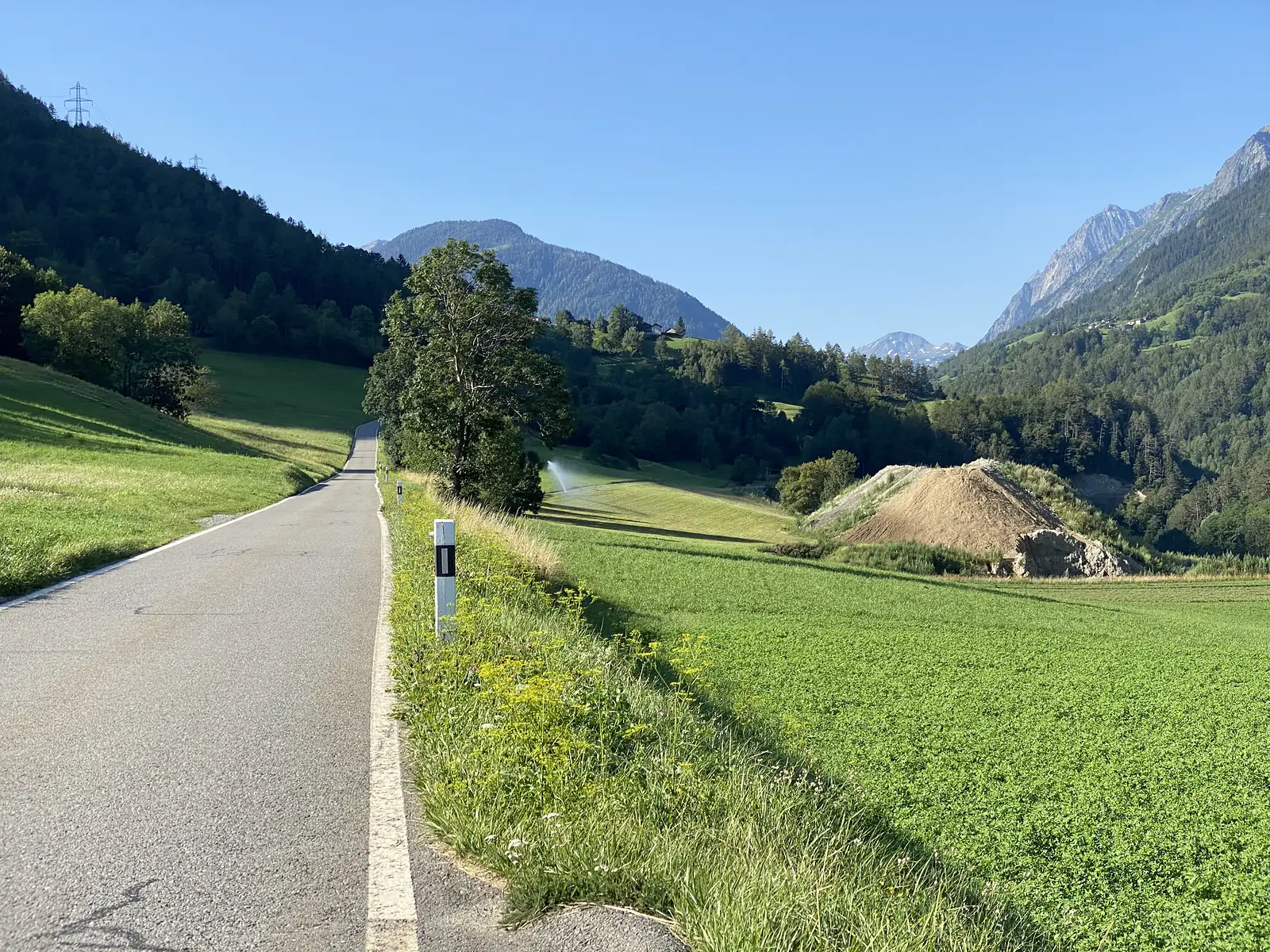 A quiet paved road running through a green valley between wooded hillsides, with a mound of excavated earth and snow-flecked peaks in the distance.
