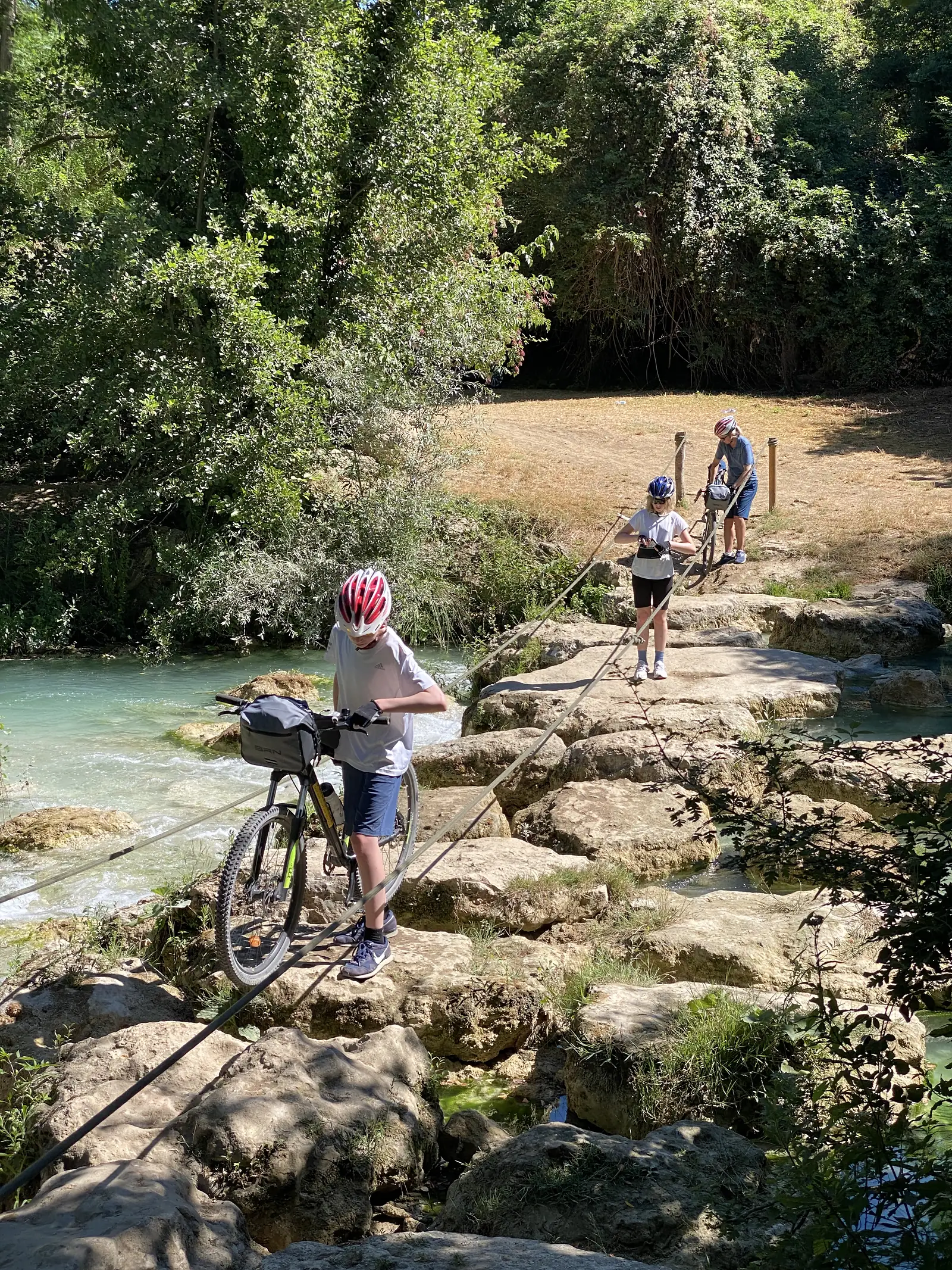 Cyclists wheel their bikes across pale travertine rock terraces beside a cascading stream, with family members walking ahead on the bank.
