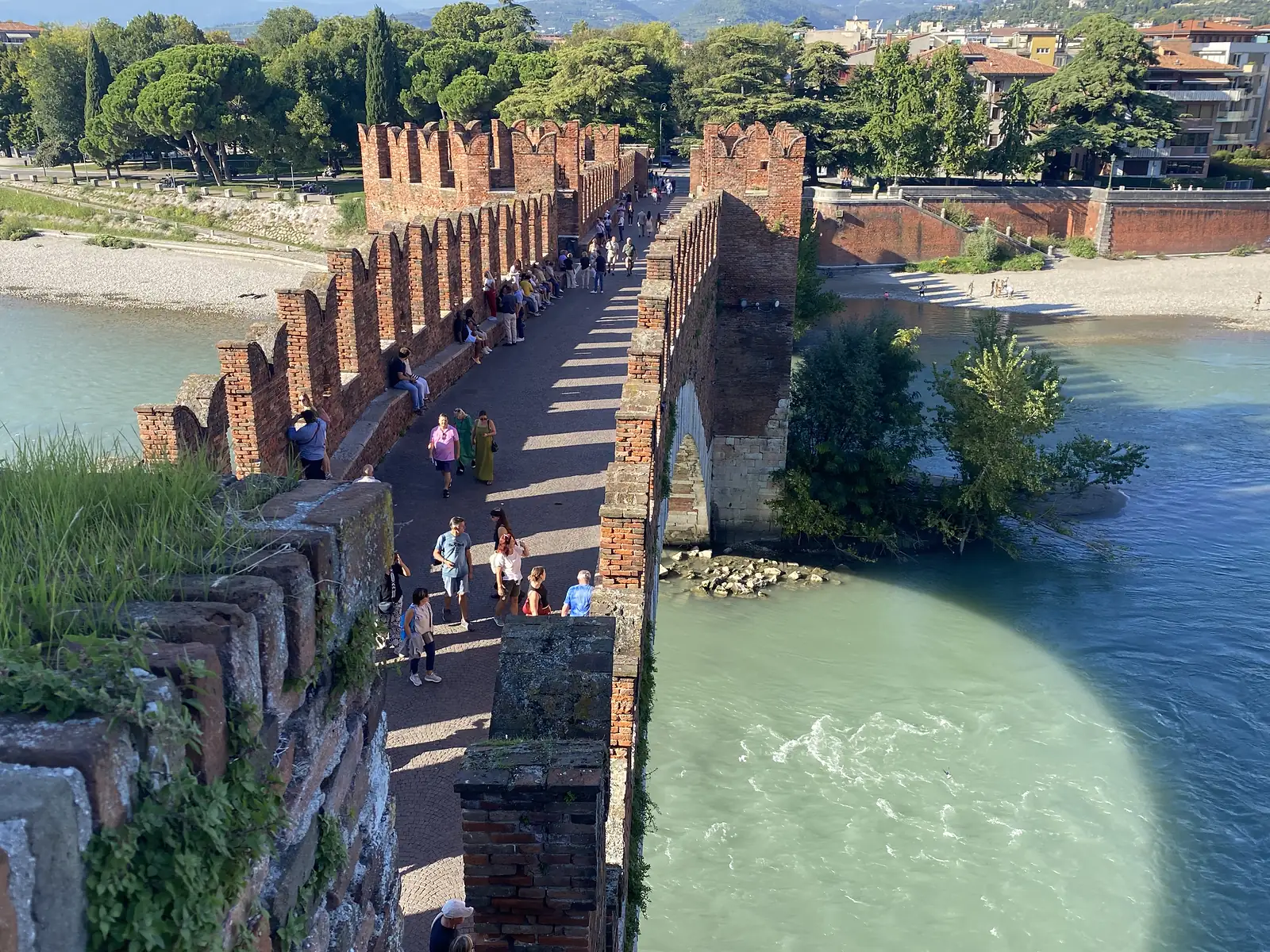 People stroll across a red-brick fortified bridge with swallowtail battlements over a green river, the town spread out beyond.