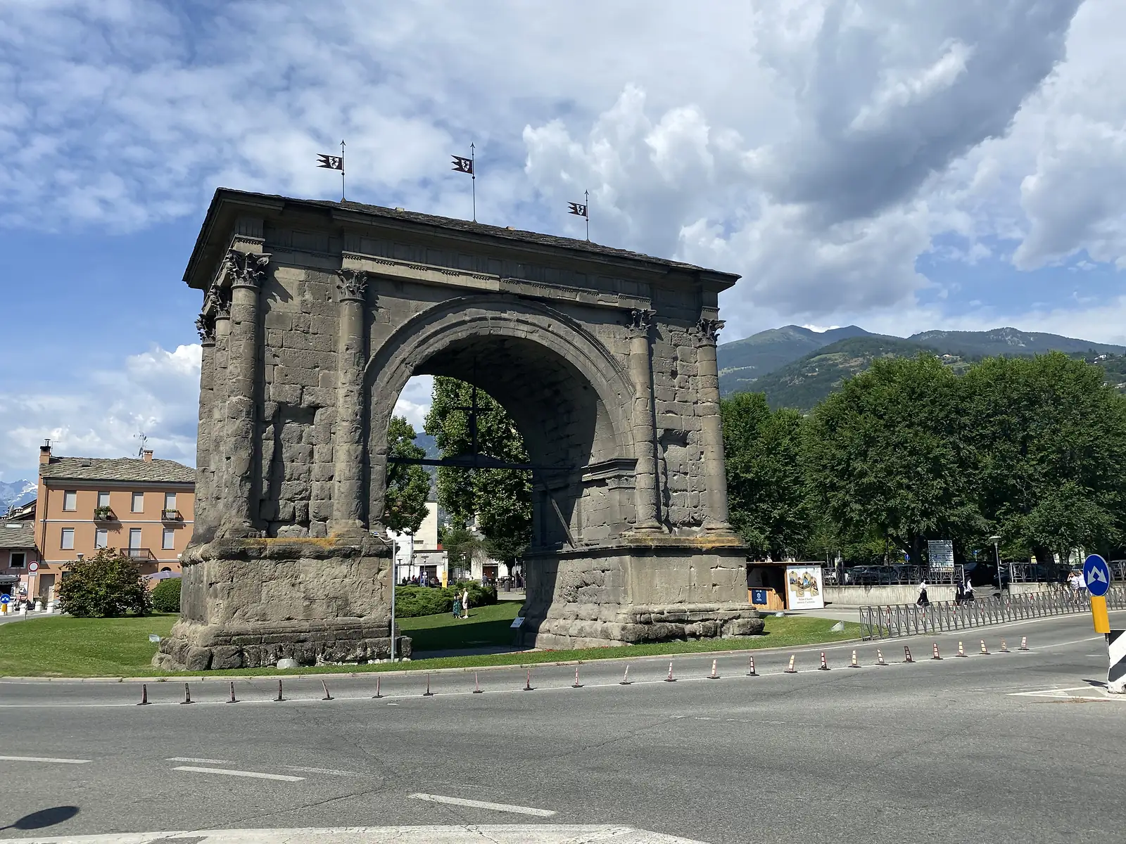 A weathered Roman-era triumphal arch with fluted columns and small pennants on its roofline stands on a traffic island, forested mountains rising behind.