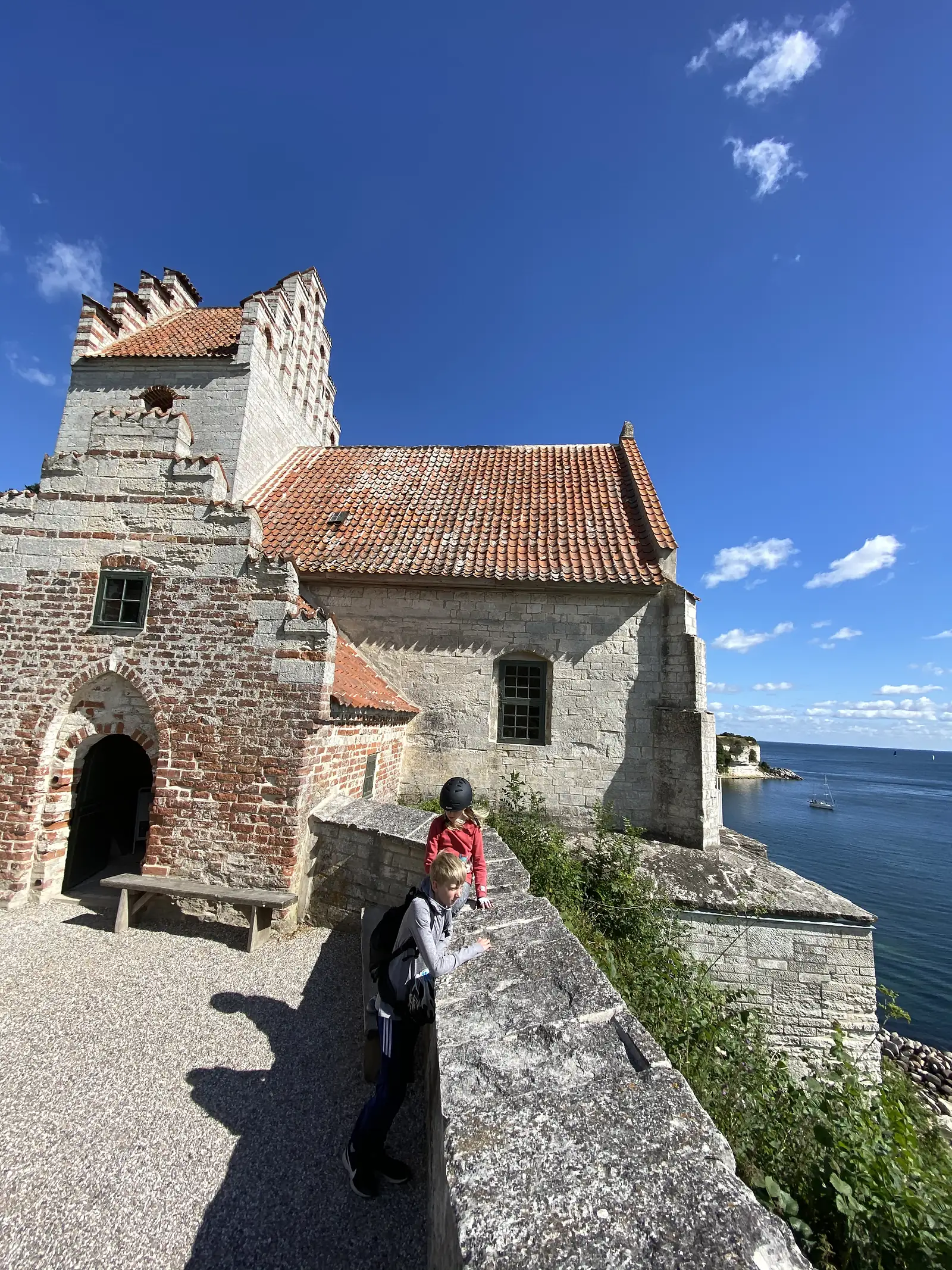 Two children leaning on a stone parapet beside a stepped-gable cliff-top church, with the sea and a yacht at anchor below.