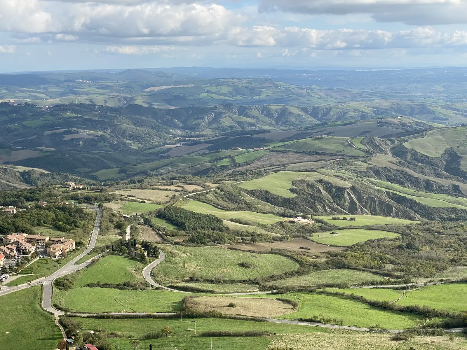 A sweeping aerial view from a hilltop over a patchwork of green fields and wooded ridges, with a small village and winding road in the foreground and layered hills receding to the horizon under a cloudy sky.